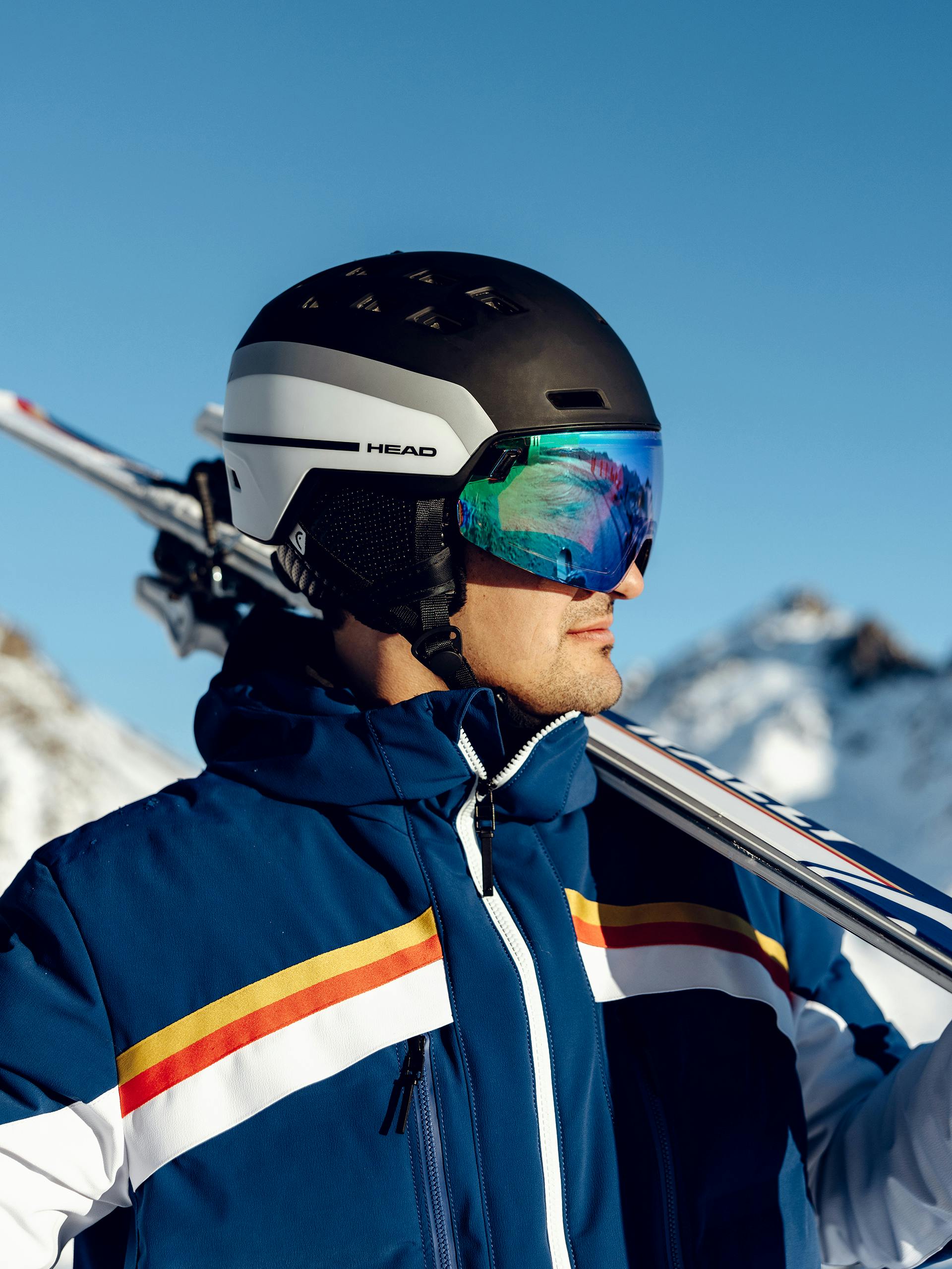 A man is pictured close up looking to the side, wearing the new Porsche Head ski helmet, Porsche ski jacket and holding Porsche skis on his shoulder.