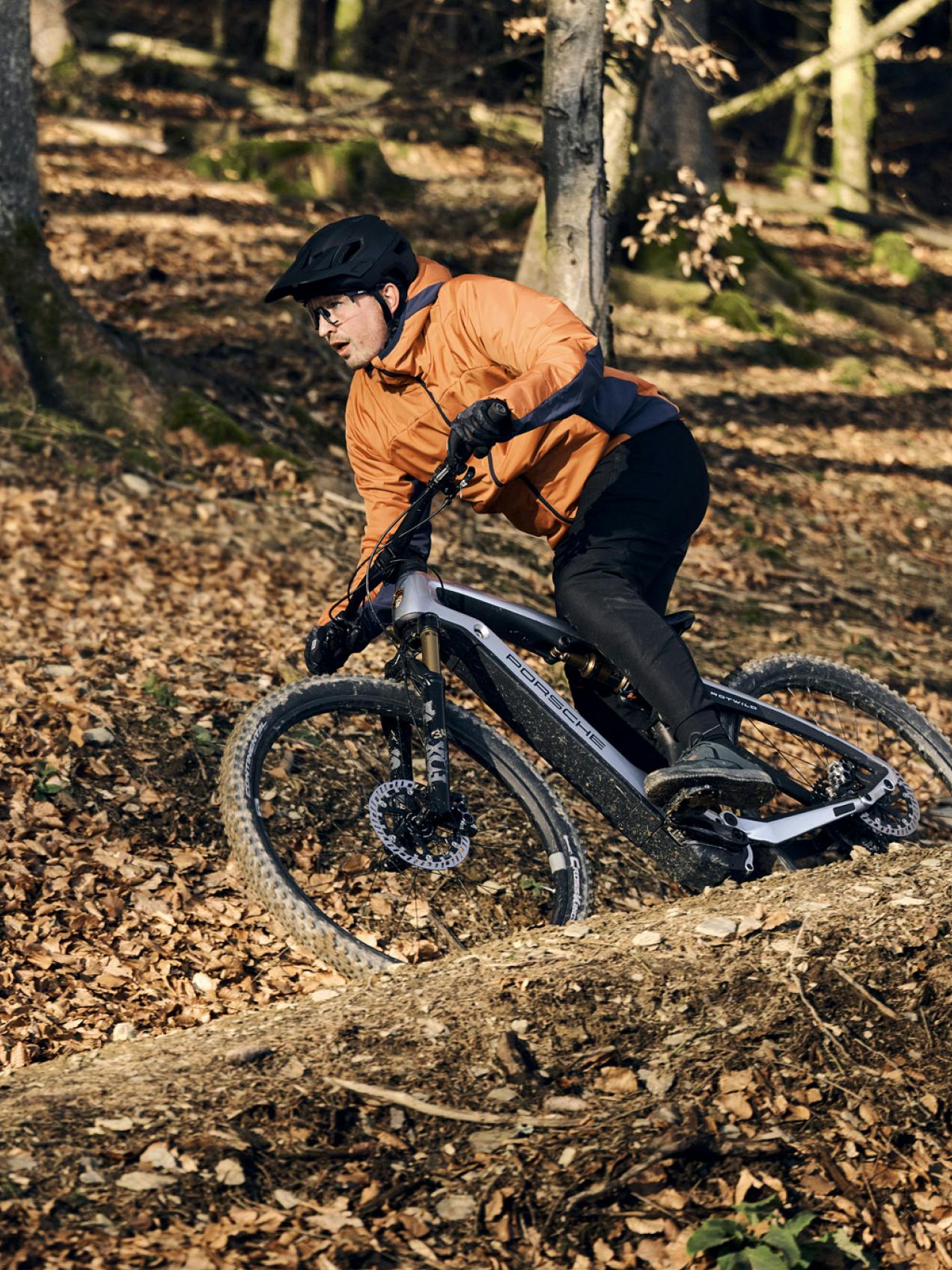 A cyclist is pictured wearing an orange out-door jacket and riding the Porsche Ebike Cross on an unpaved path.