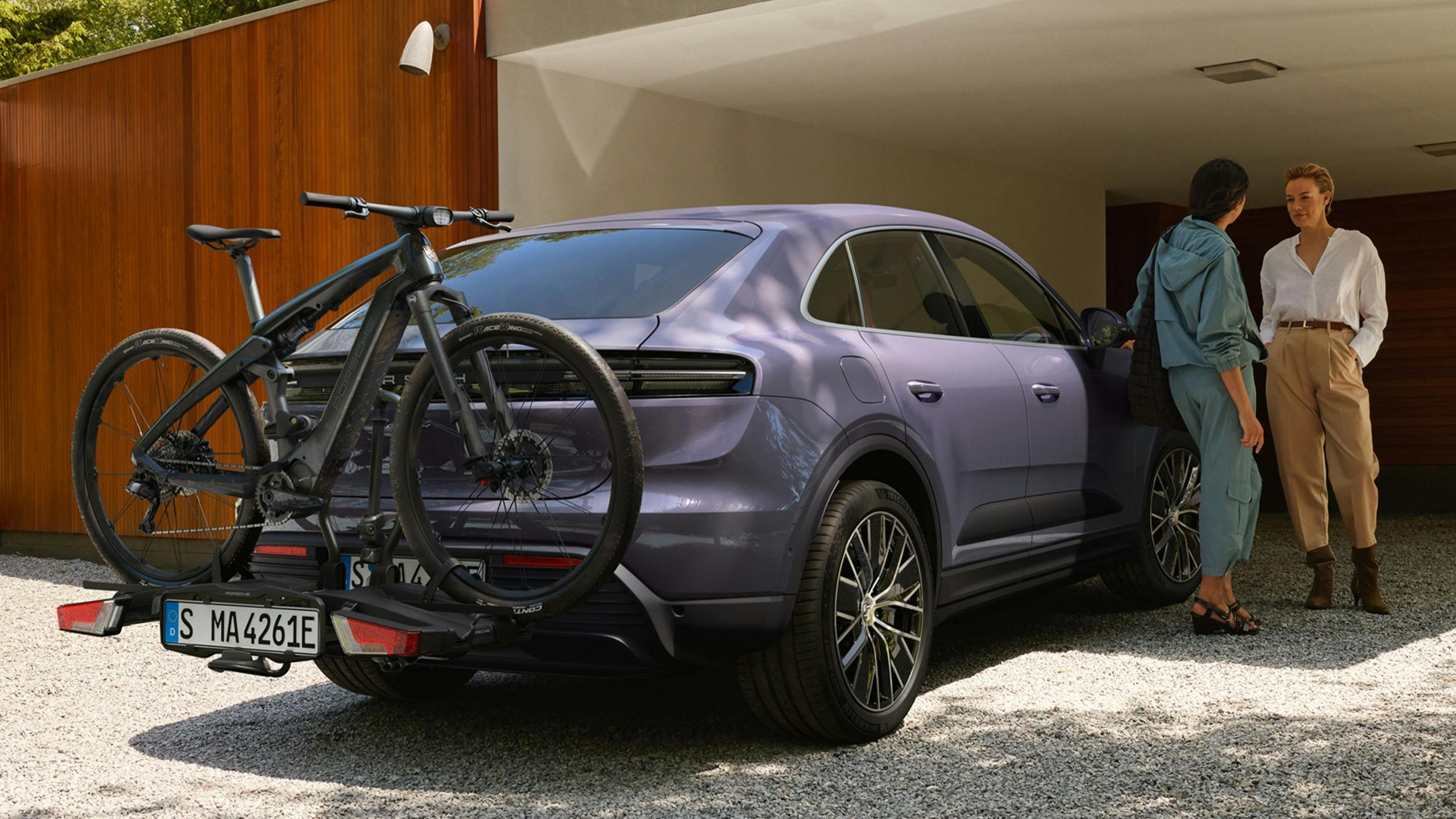 Two women chatting, standing next to a Provence coloured electric Macan with a Porsche eBike on a bike rack attached to the back.