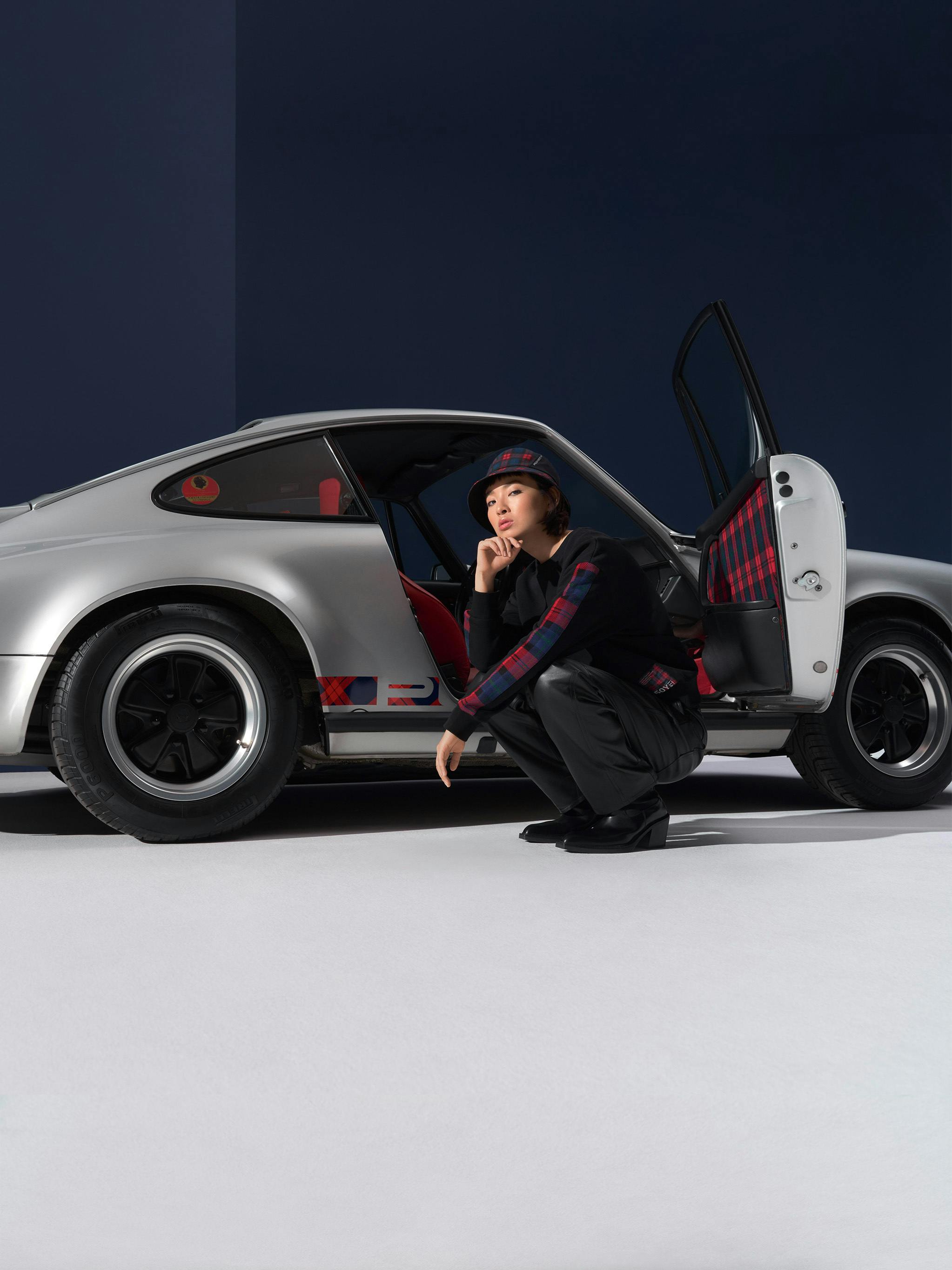 Woman sitting next to a silver Porsche 911 wearing Porsche Turbo No. 1 collection clothing