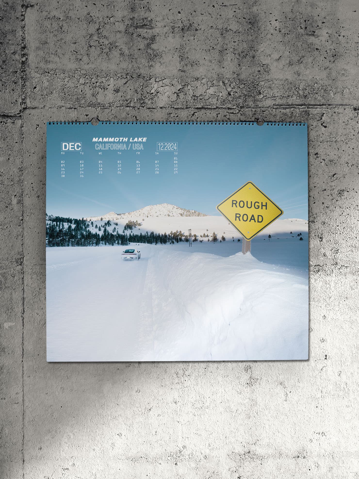 Pictured is a Porsche wall calendar for the year 2024 on a gray concrete wall. The calendar shows a white snowy landscape with a white Porsche 911 and a yellow sign with the inscription "Rough Road".