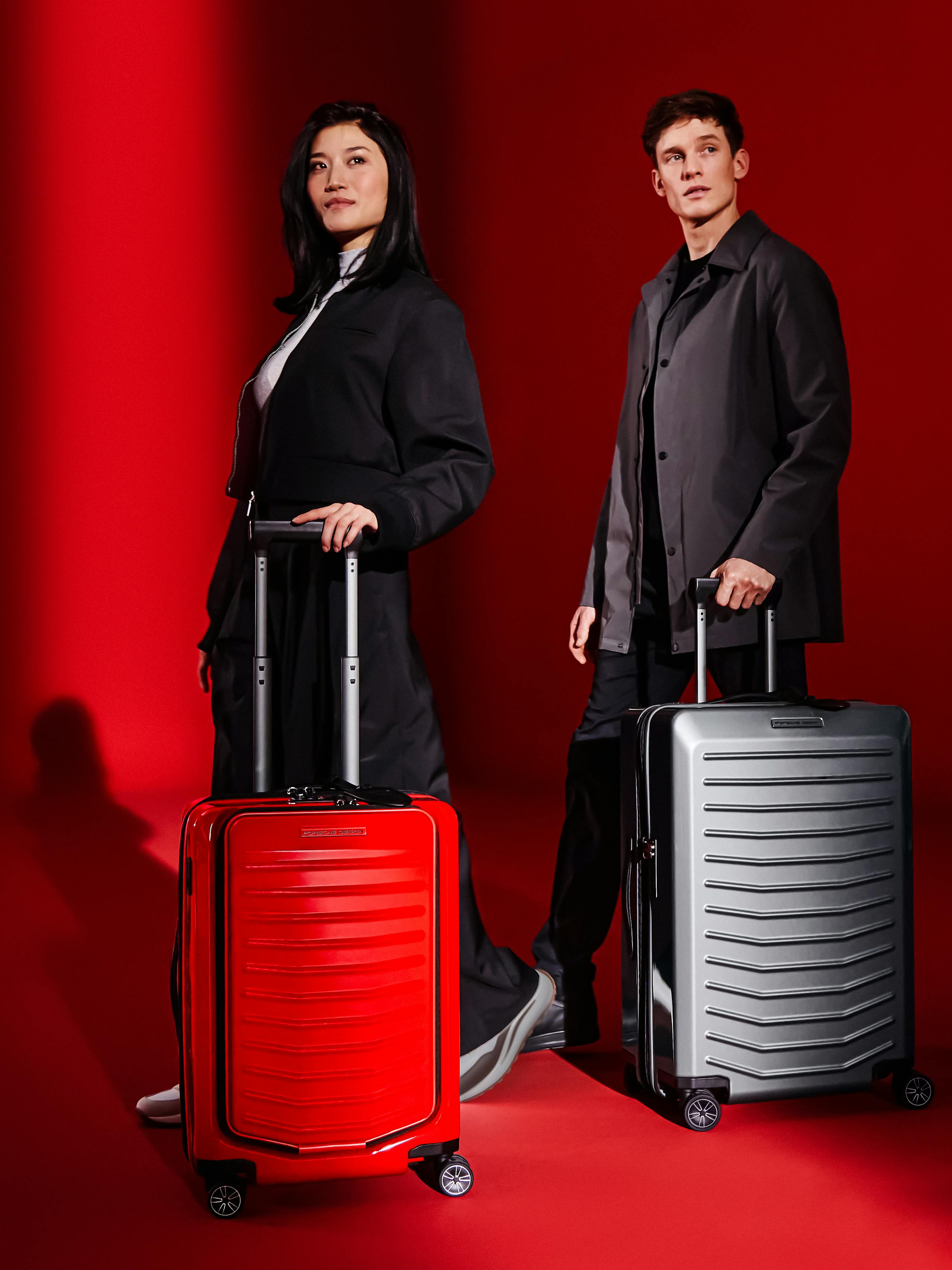 A woman pushing a red Porsche Design hardcase trolley suitcase, in front of a man pushing a grey Porsche Design Hardcase Trolley suitcase.