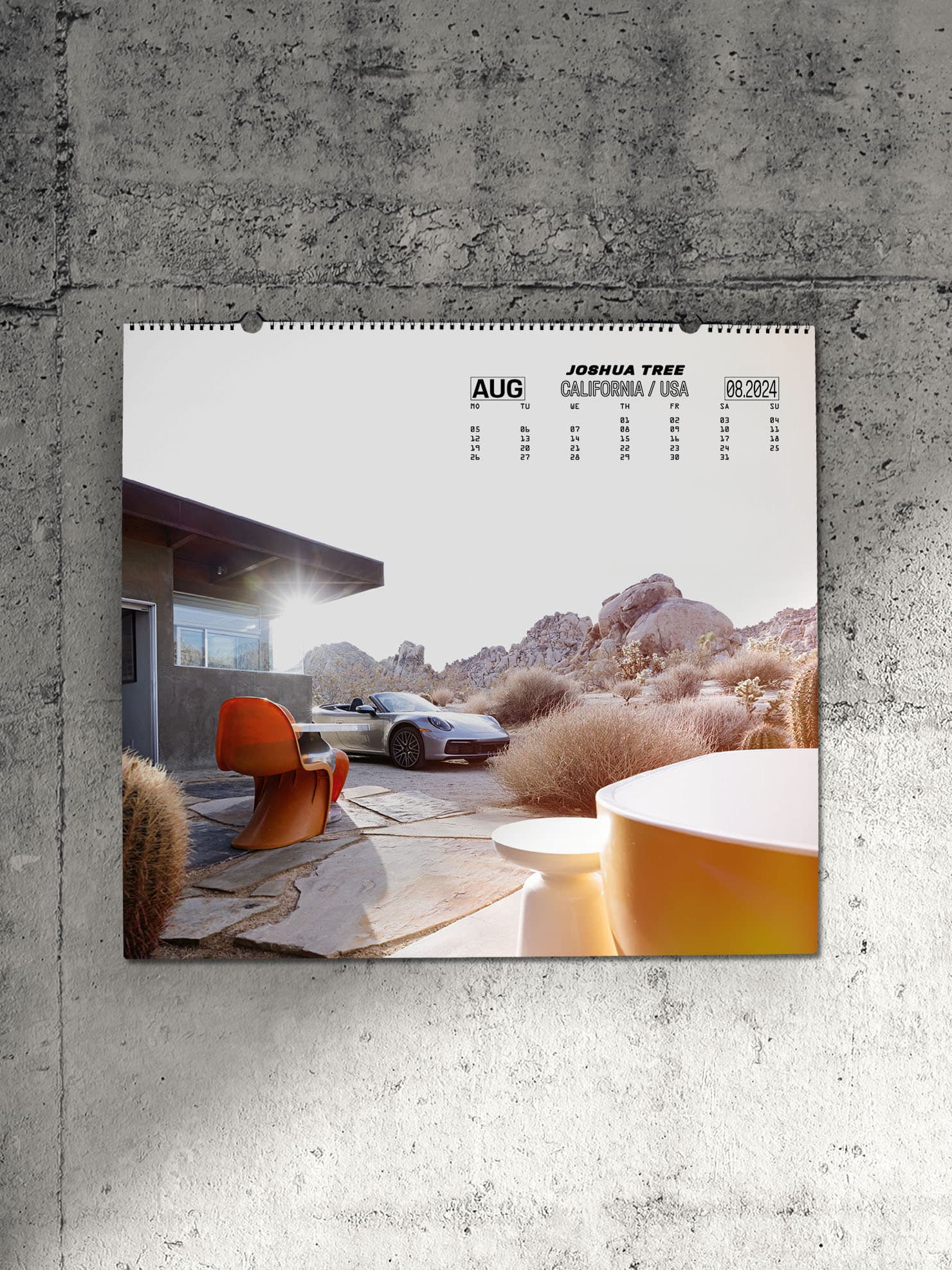 Pictured is a Porsche wall calendar for the year 2024 on a gray concrete wall. The calendar shows a silver Porsche 911 with a house in a desert landscape.