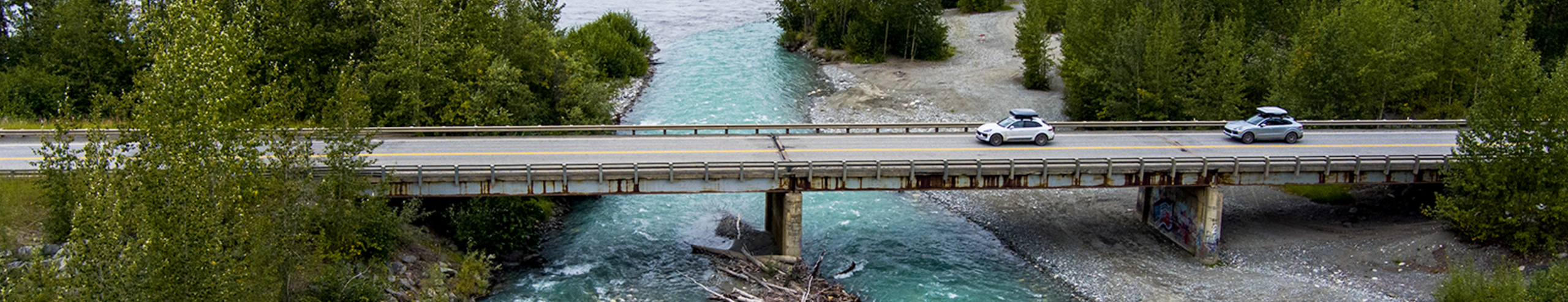 You can see two Porsches with a roof box driving on a bridge over a river 