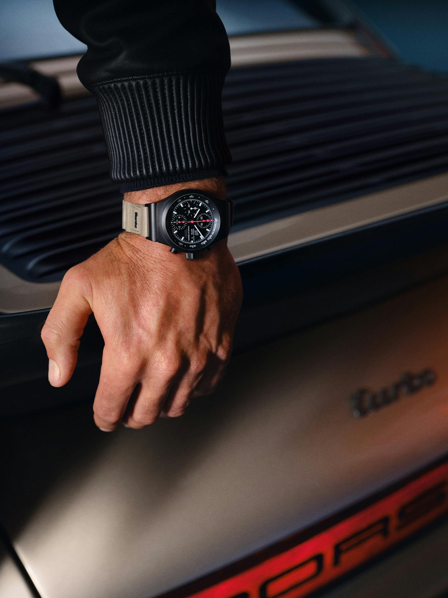 Man is leaning on the back of the car and wearing Porsche Design 50Y Chronograph