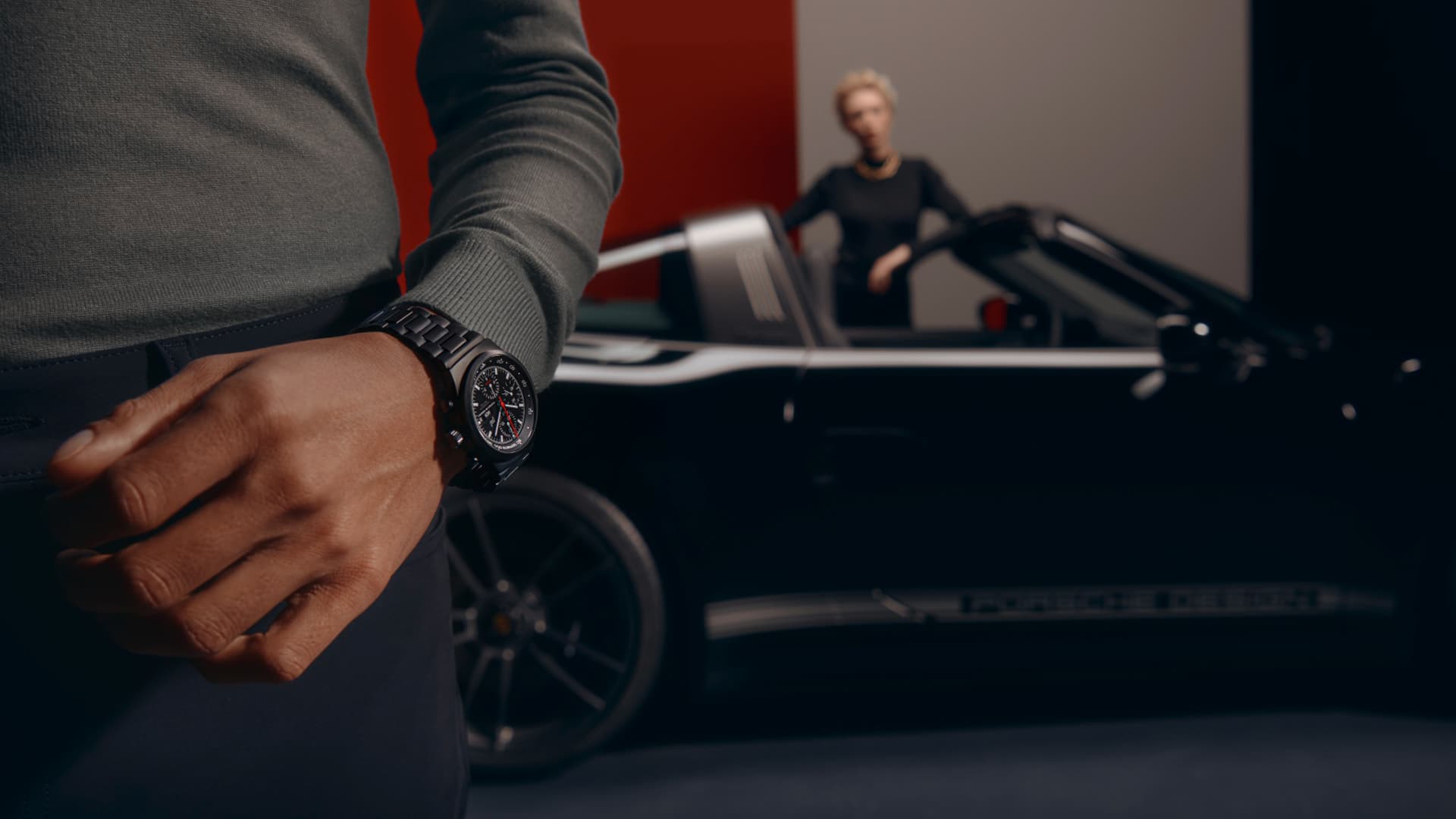Mężczyzna w czarnym zegarku na rękę Porsche; w tle czarne Porsche 911 z mężczyzną w środku.