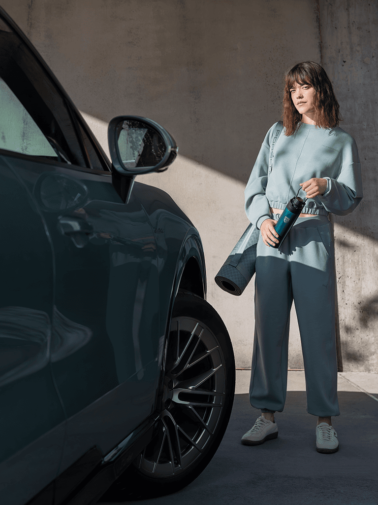 Femme en survêtement bleu tenant une bouteille d'eau et un tapis de yoga, debout près d'une voiture bleue au soleil.