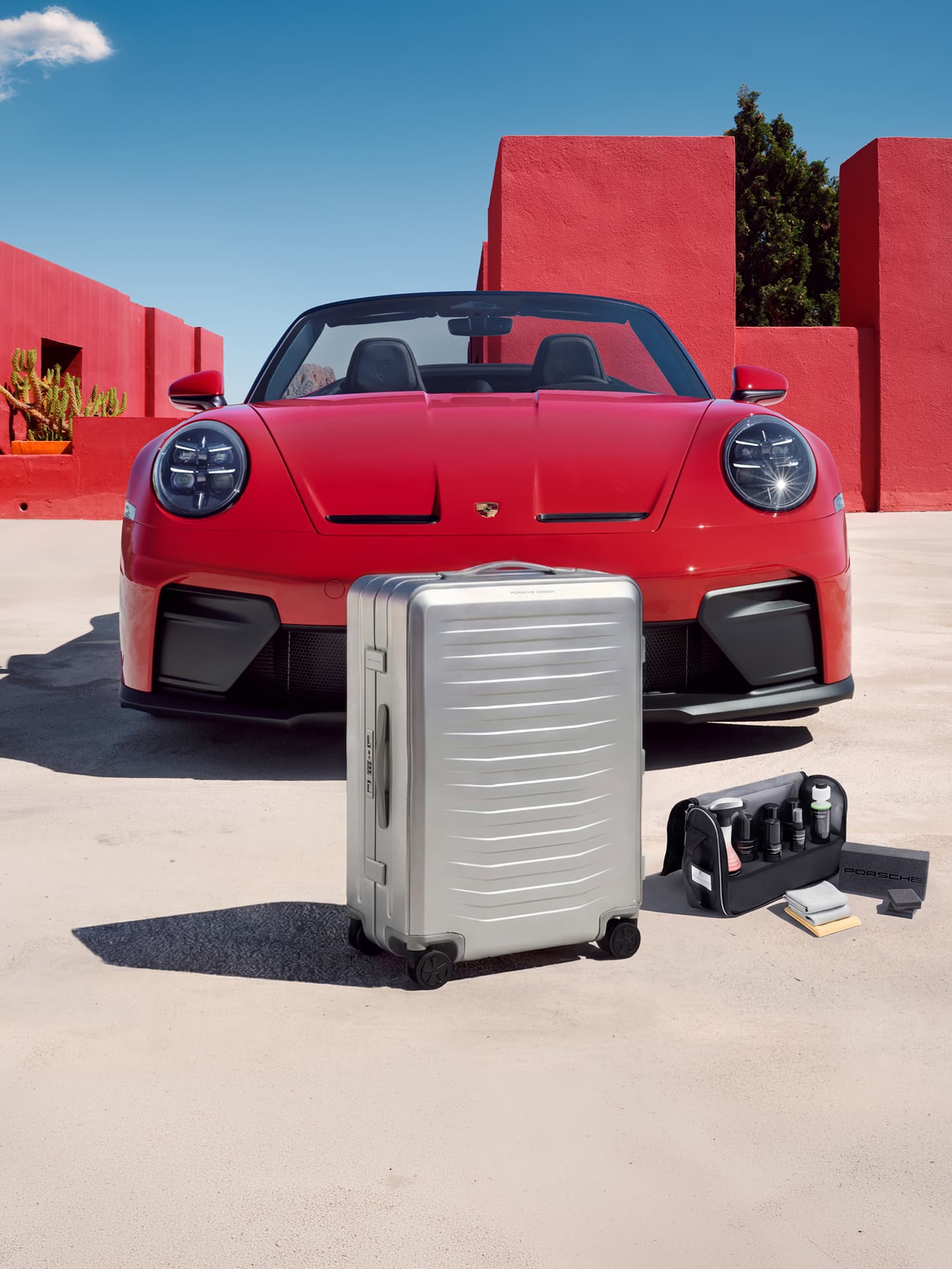Valigia rigida argentata e set per la cura davanti a una Porsche Cabriolet rossa, davanti a pareti rosse sotto un cielo blu.