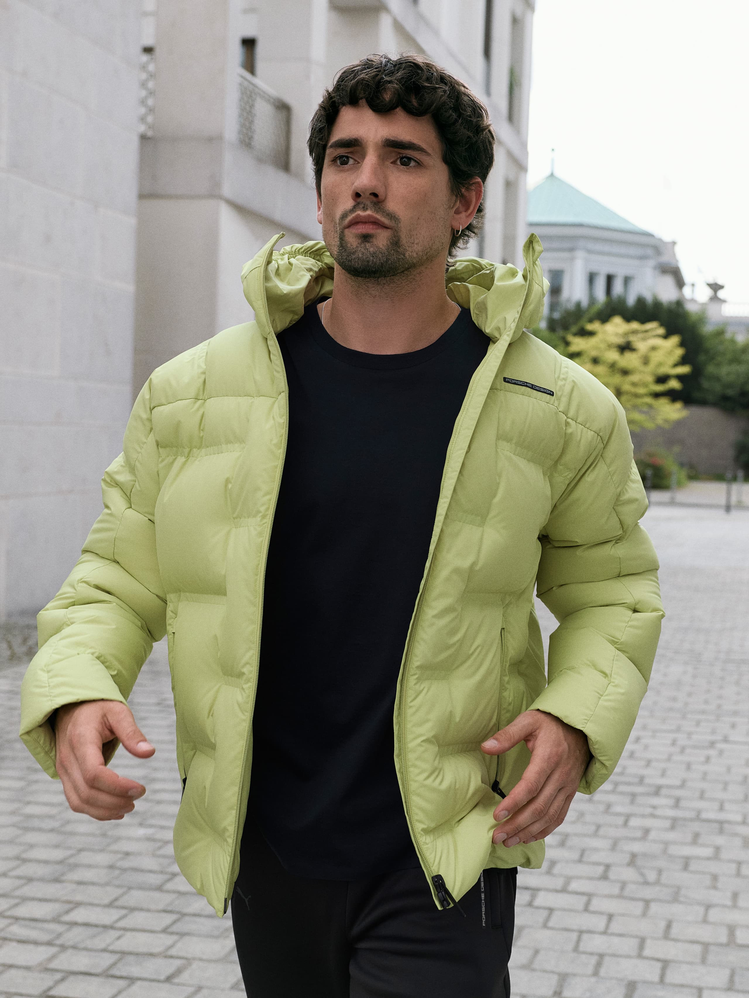 Man in a light green puffer jacket walking on a cobblestone path, with a modern building and trees in the background.