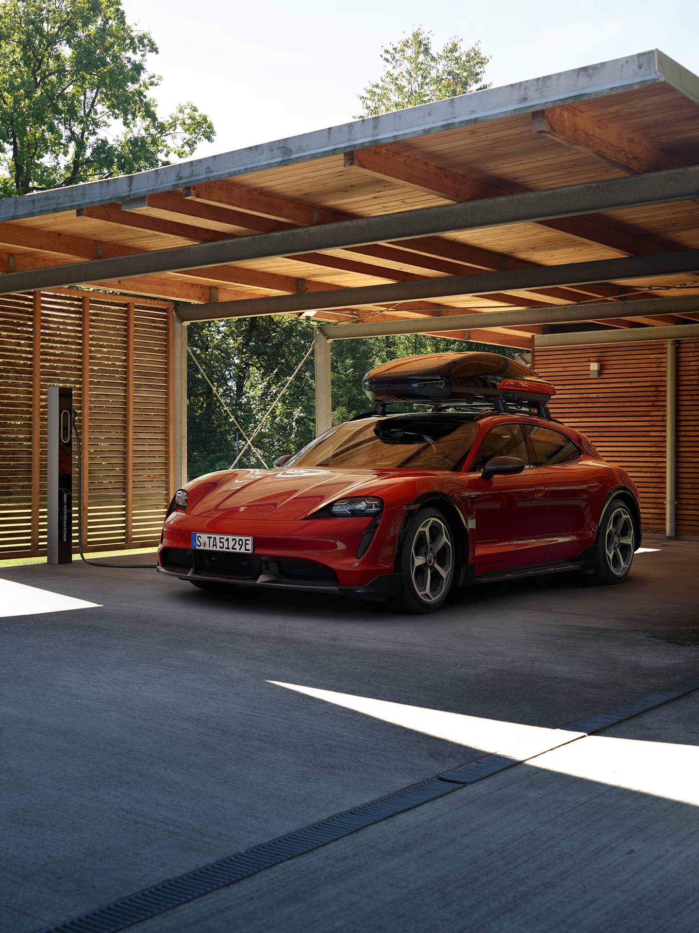 Roter Porsche Taycan Sport Turismo mit Dachbox unter modernem Carport aus Holz und Beton, im Sonnenlicht.