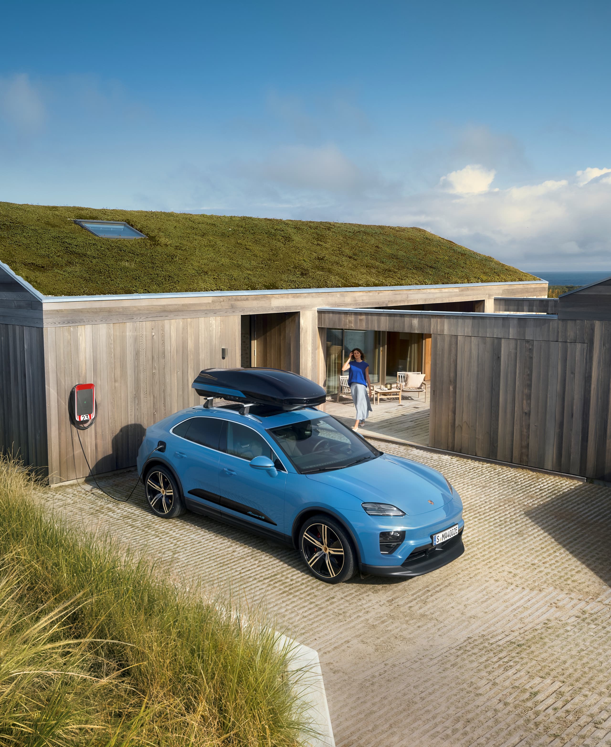 Blue Porsche SUV with roof box at a charging station in front of a modern wooden house with a green roof, person on the terrace.