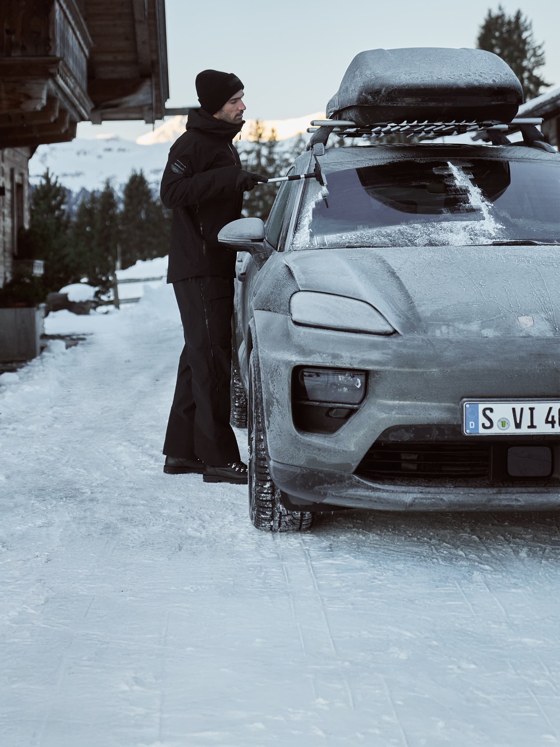 Mann in Winterkleidung kratzt Eis von der Windschutzscheibe eines grauen Porsche-SUVs, verschneite Bergkulisse, Dachbox oben.