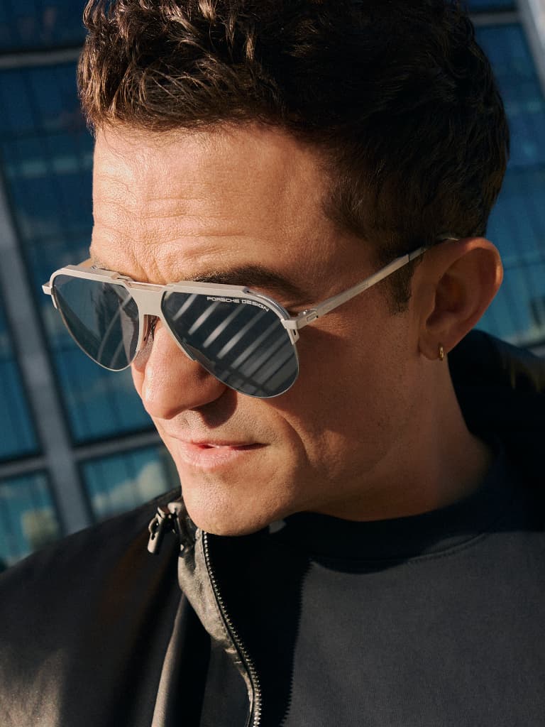 Homem a usar óculos de sol Porsche Design prateados com lentes escuras, em primeiro plano, em frente a um edifício moderno de vidro.