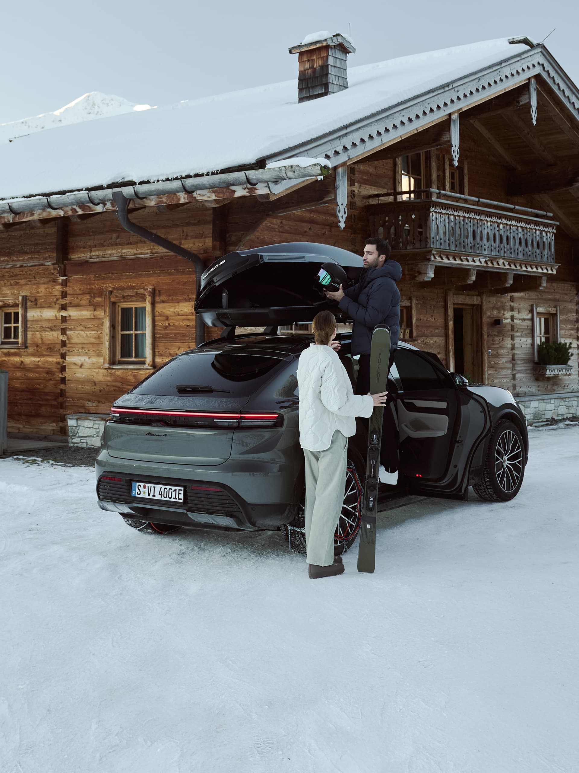 Man and woman loading skis into a black Porsche near a snowy cabin, with mountains in the background.
