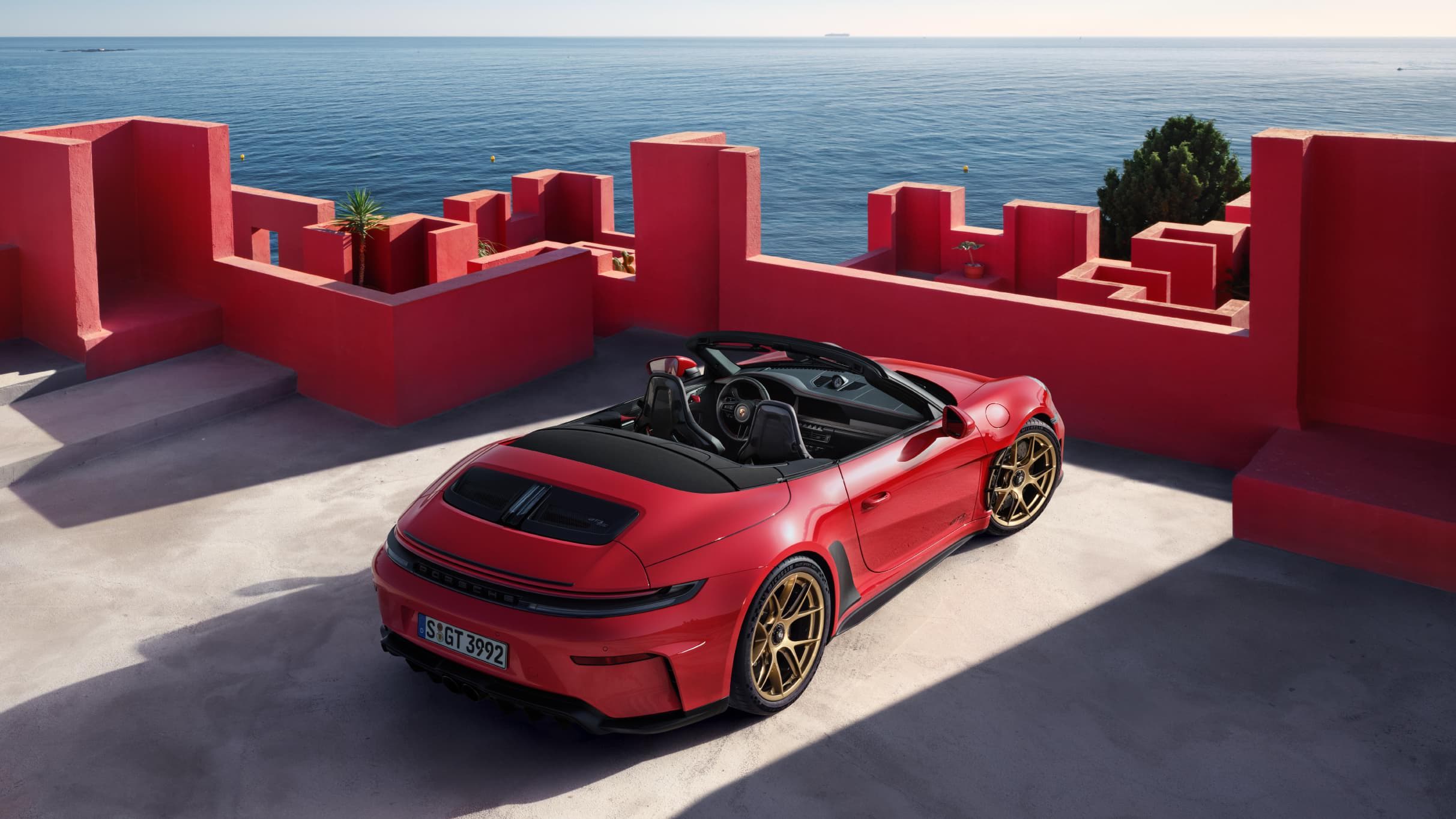 Cabriolet Porsche 911 rouge avec jantes dorées, garé devant des murs rouges avec vue sur la mer.