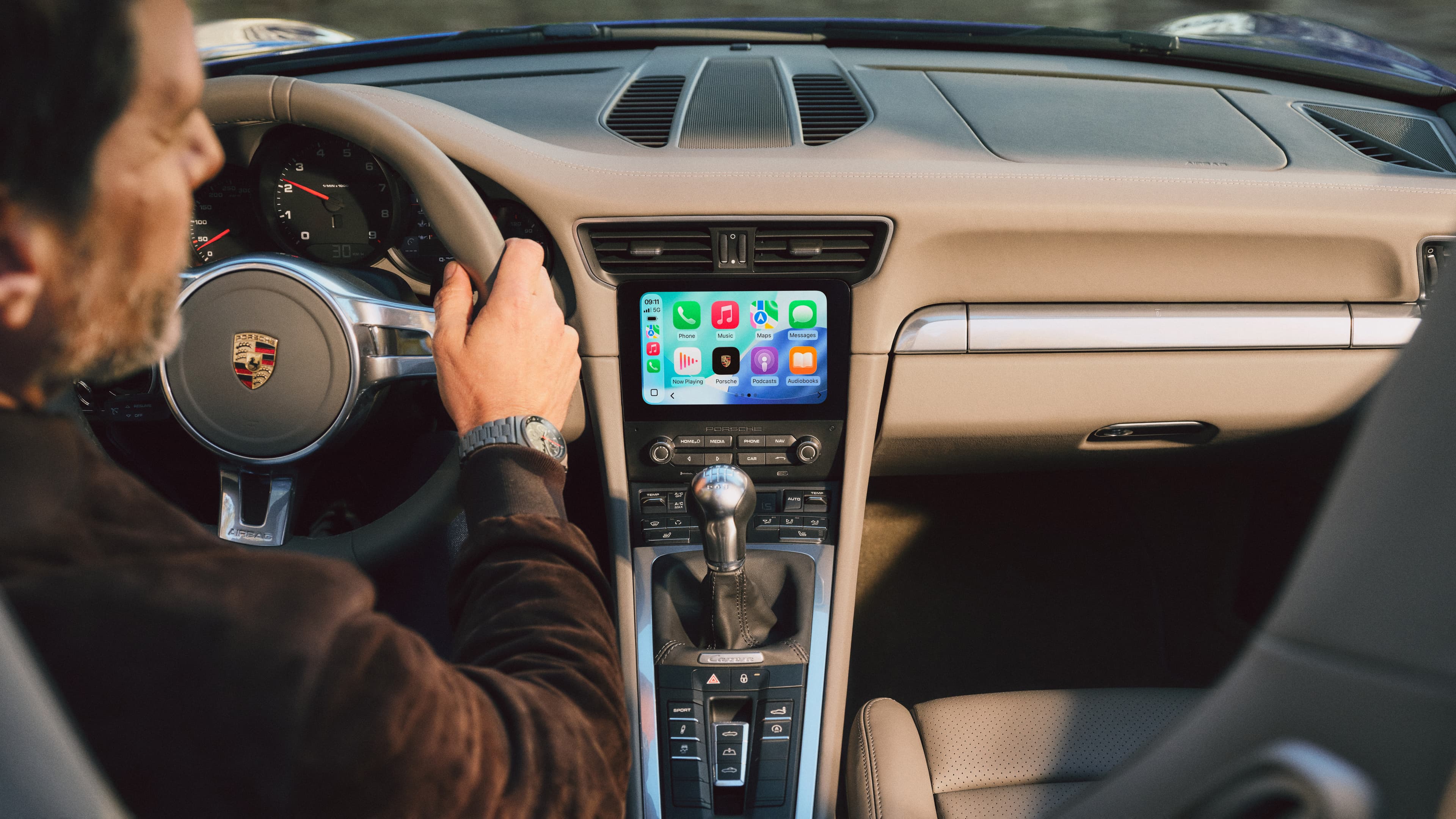 Kierowca w kokpicie Porsche z beżową deską rozdzielczą; na ekranie systemu multimedialnego widoczny ekran główny Apple CarPlay.
