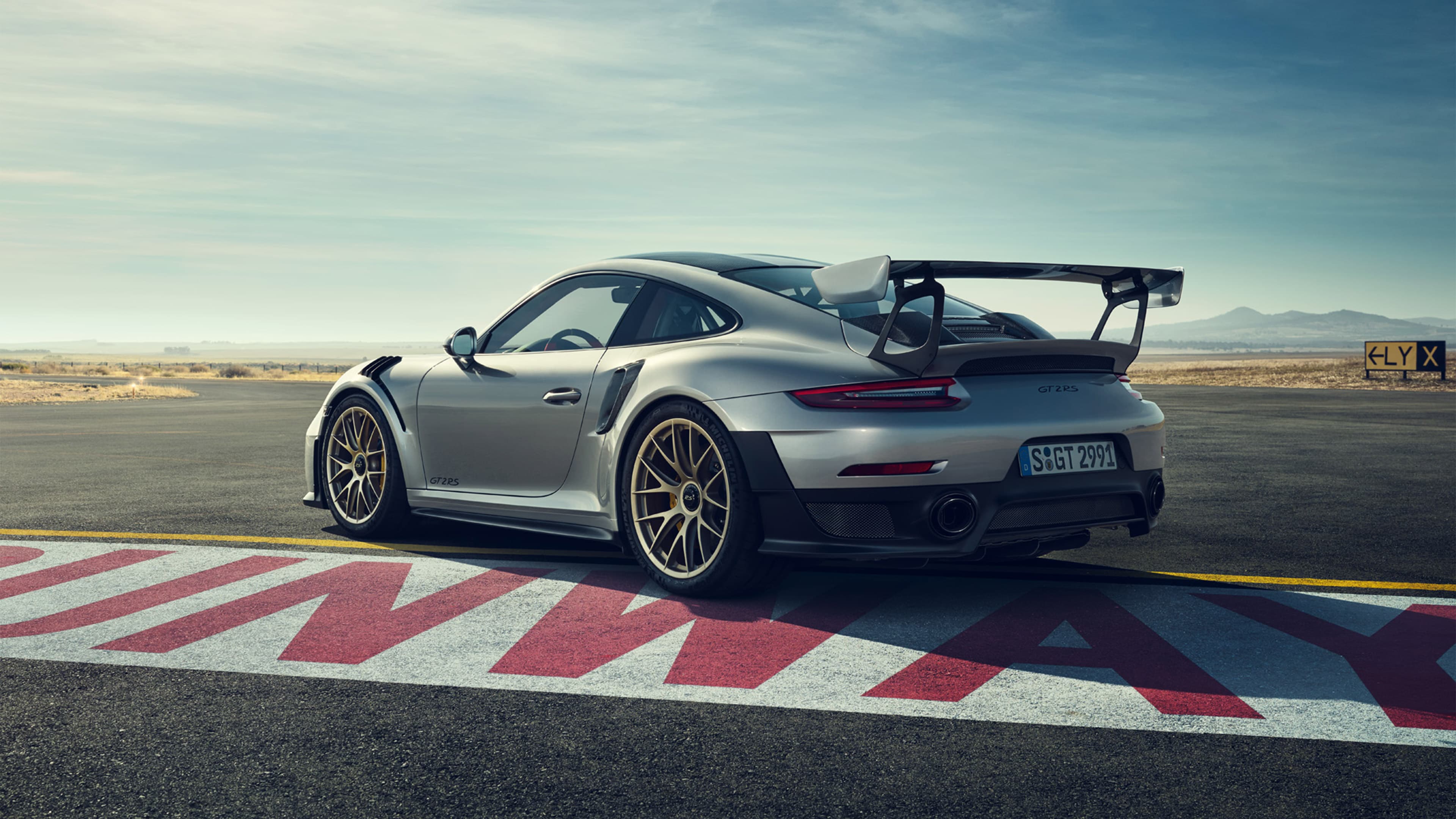 Silberner Porsche 911 GT2 RS mit großem Heckflügel auf Rennstrecke, Blick von hinten links, weiter Himmel.
