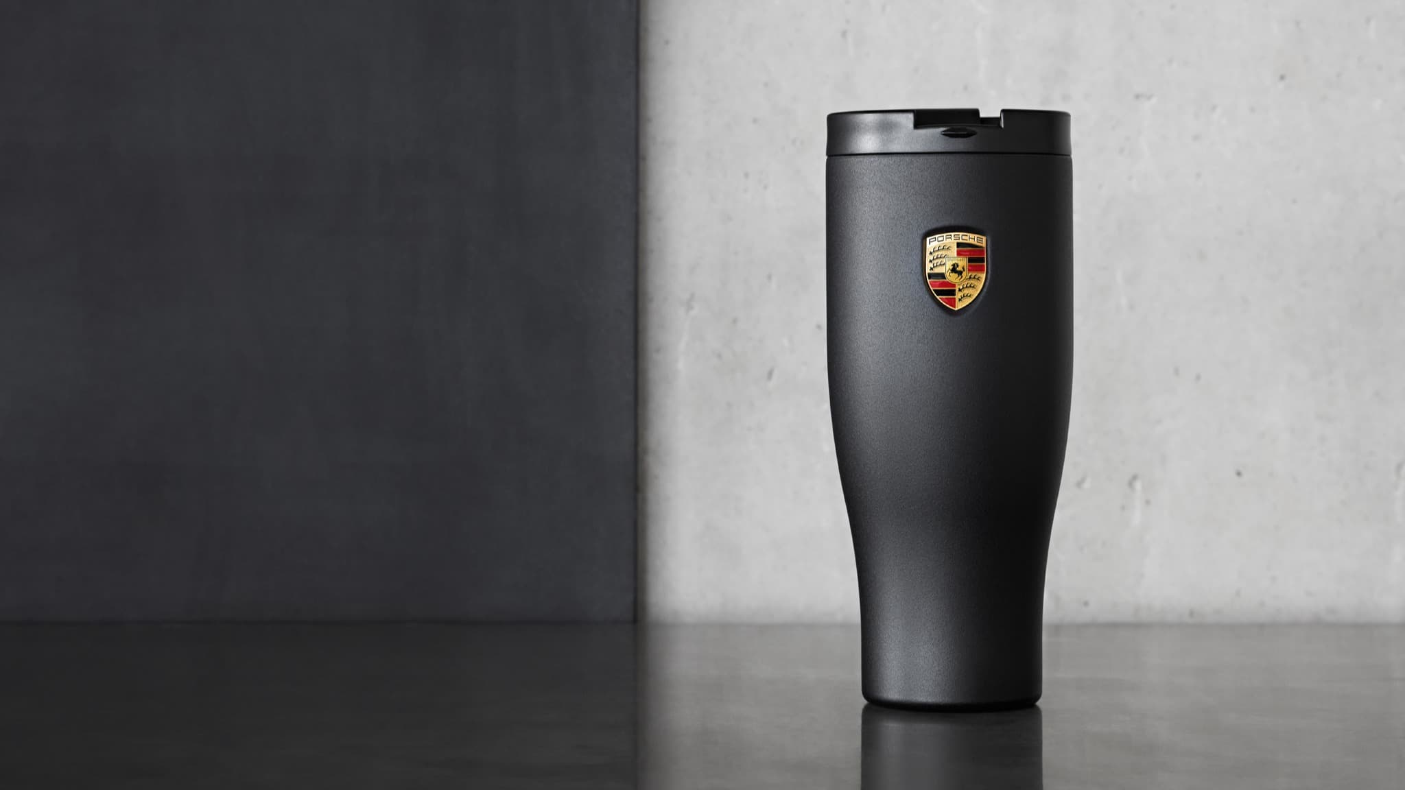 Black Porsche travel mug with logo, placed on a dark surface against a gray background.