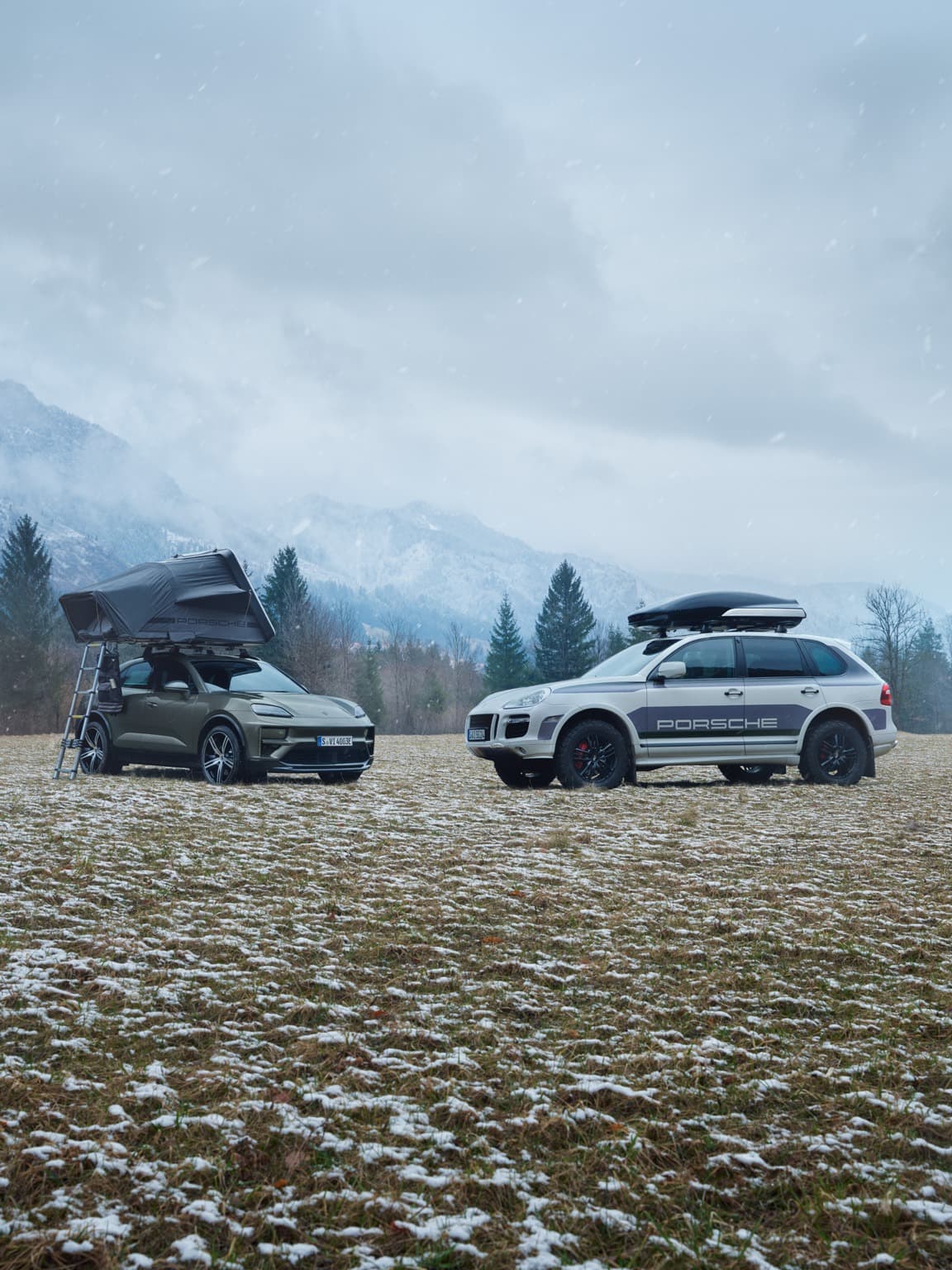 Zwei Porsche SUVs mit Dachzelten auf einem verschneiten Feld, umgeben von Bergen und Bäumen unter bewölktem Himmel.
