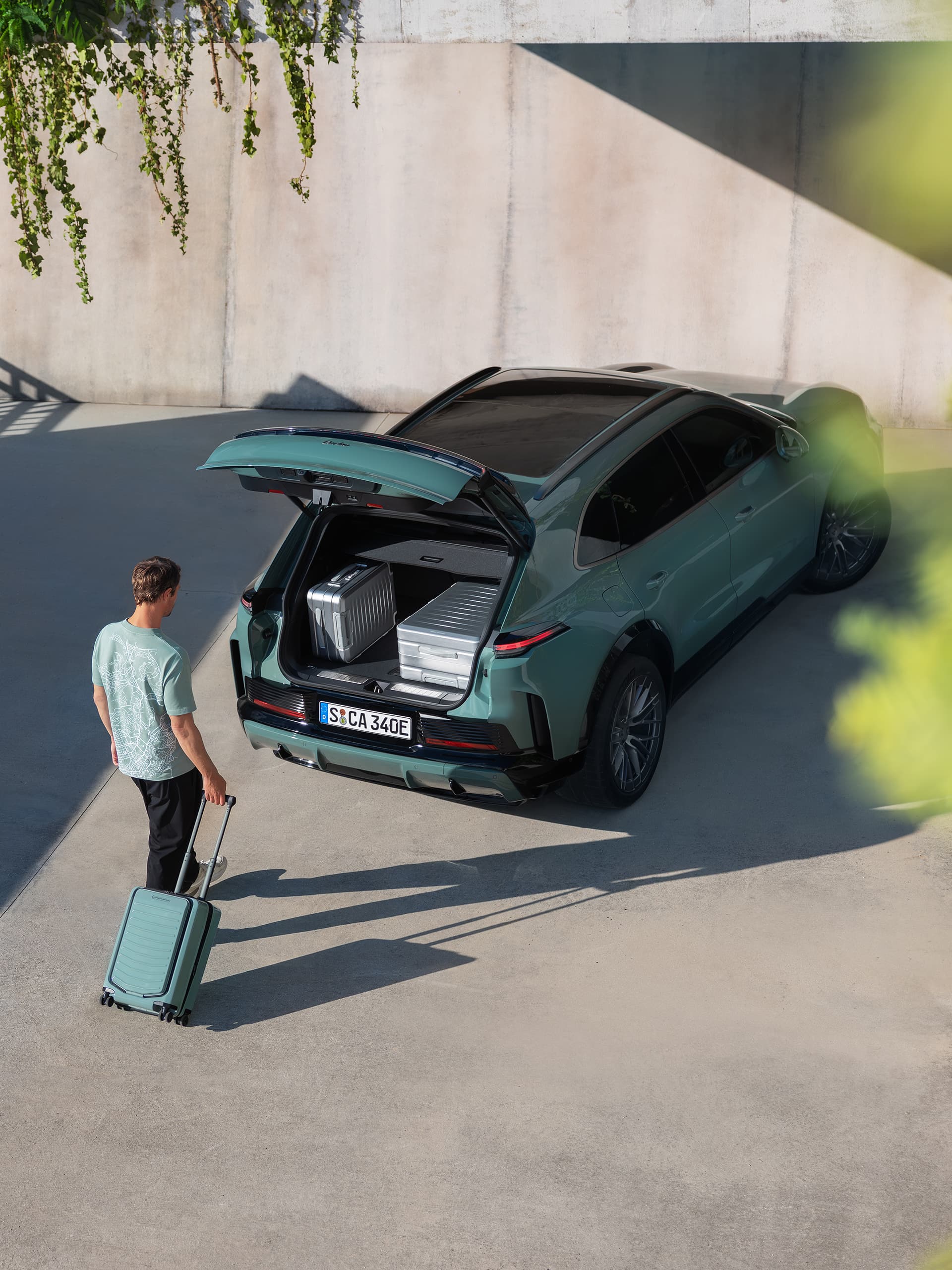 Man with a turquoise suitcase stands at the open trunk of a green Porsche, luggage in the cargo area.