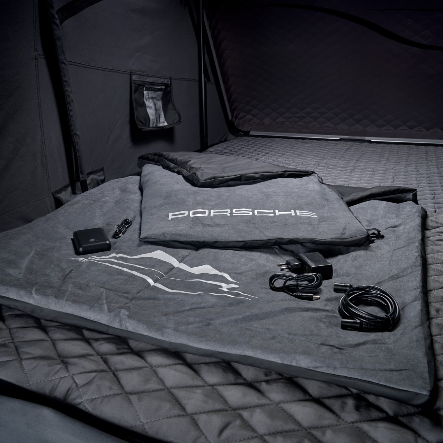 Porsche-branded gray sleeping bag with charger and cables on quilted surface inside a dark tent.