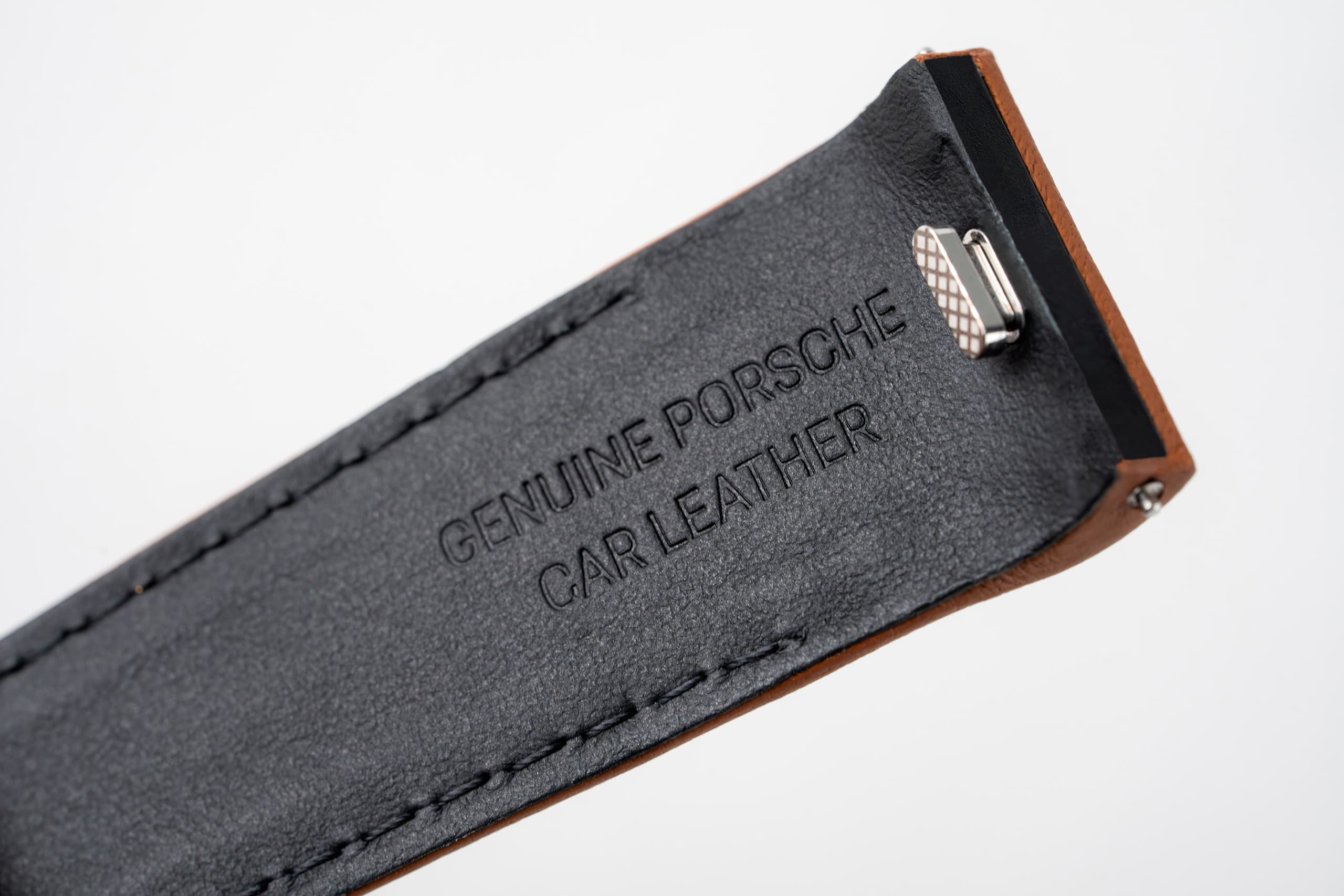 Close-up van een zwarte leren riem met reliëftekst “Genuine Porsche Car Leather” en een metalen clip.