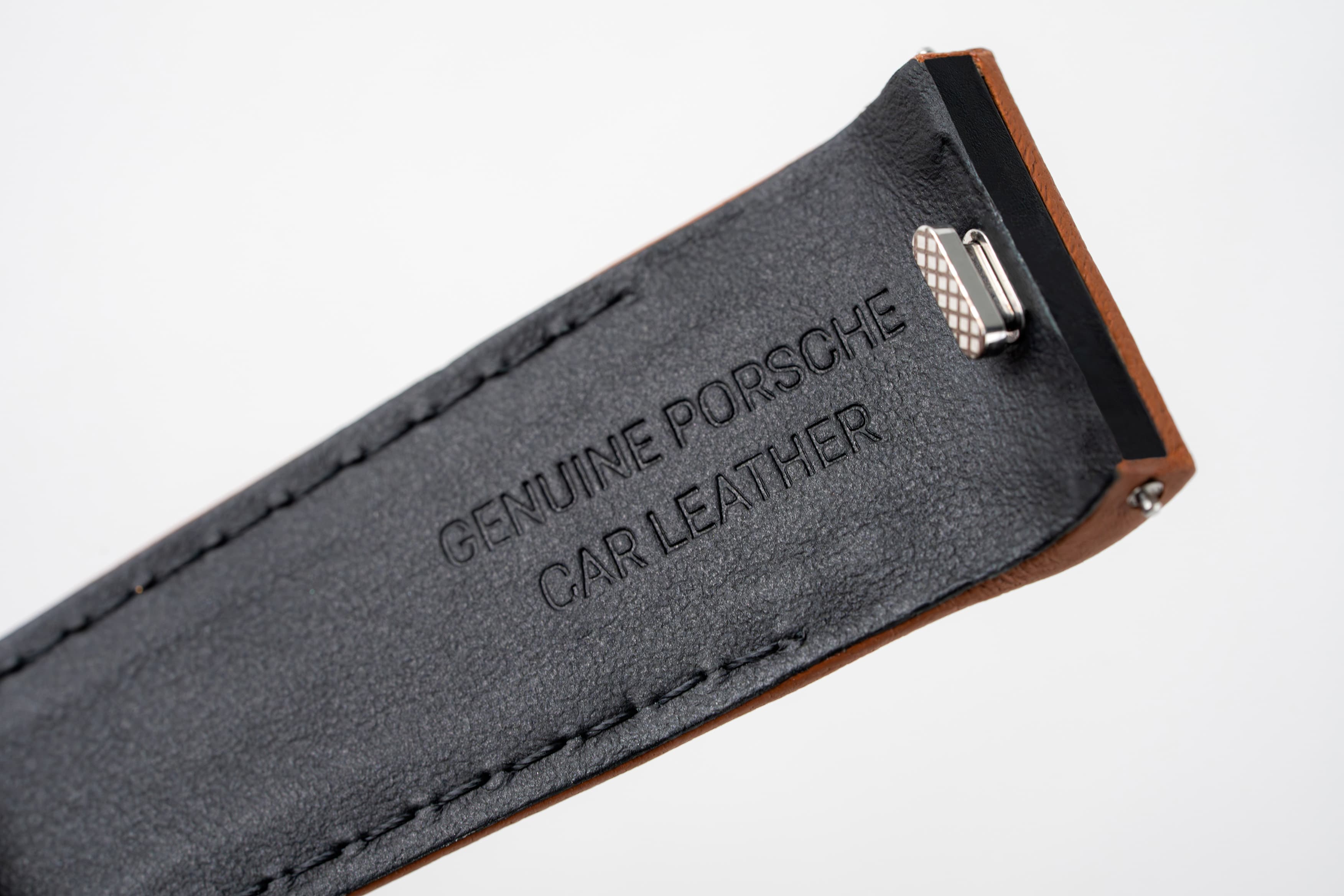 Primer plano de una correa de cuero negro con el grabado «Genuine Porsche Car Leather» y un clip metálico.