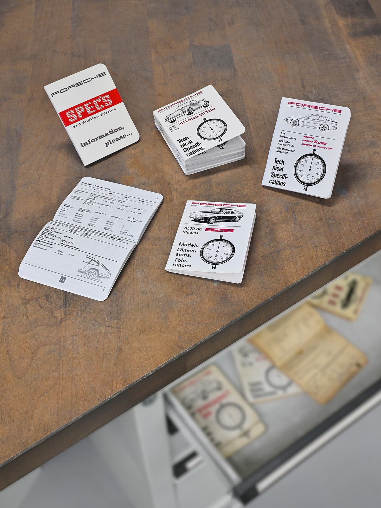 Porsche spec booklets on a wooden table, featuring technical details and car illustrations, with some open and others stacked.
