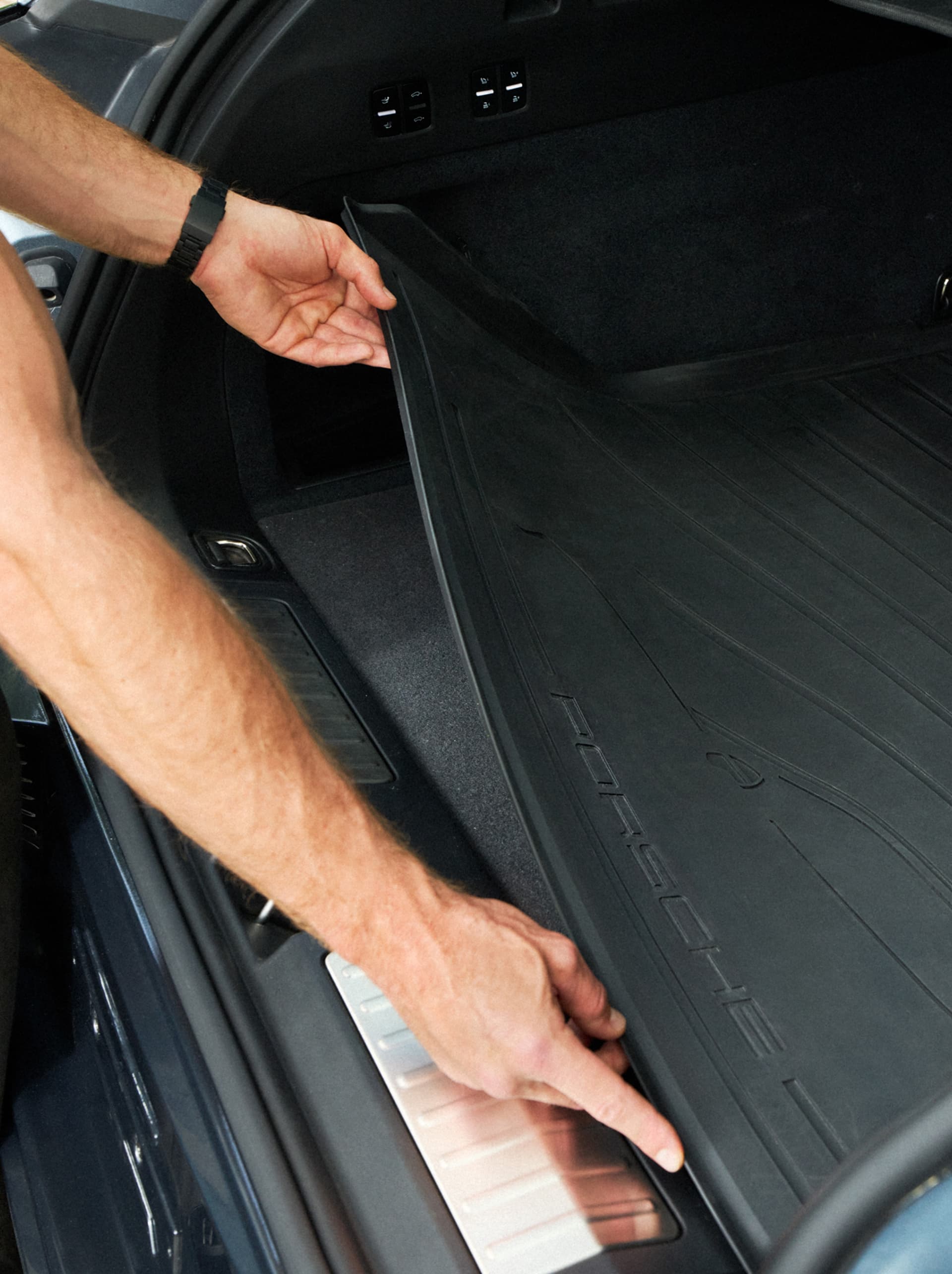 Hand placing black Porsche cargo liner into the rear trunk, close-up of textured rubber mat and sill plate.