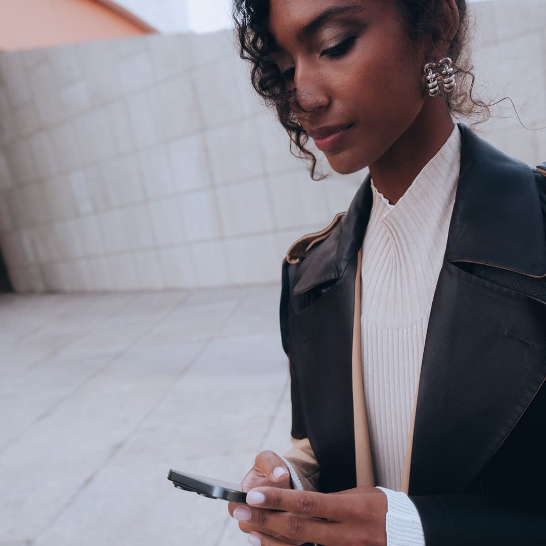 Woman in a black leather jacket and white top, looking at a smartphone, standing against a tiled wall background.