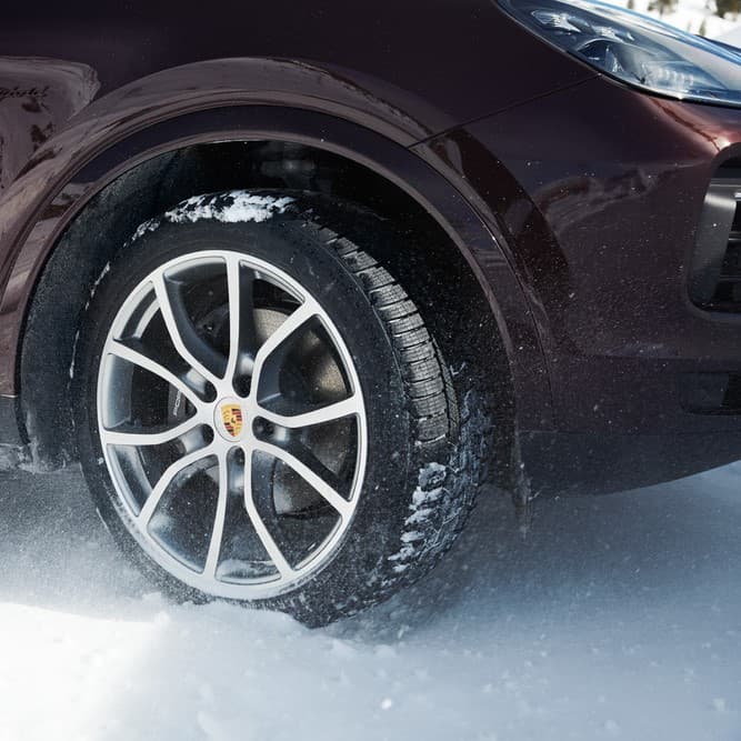Nahaufnahme eines Porsche-Rads auf schneebedecktem Untergrund, mit silberner Alufelge und dunkelroter Karosserie.