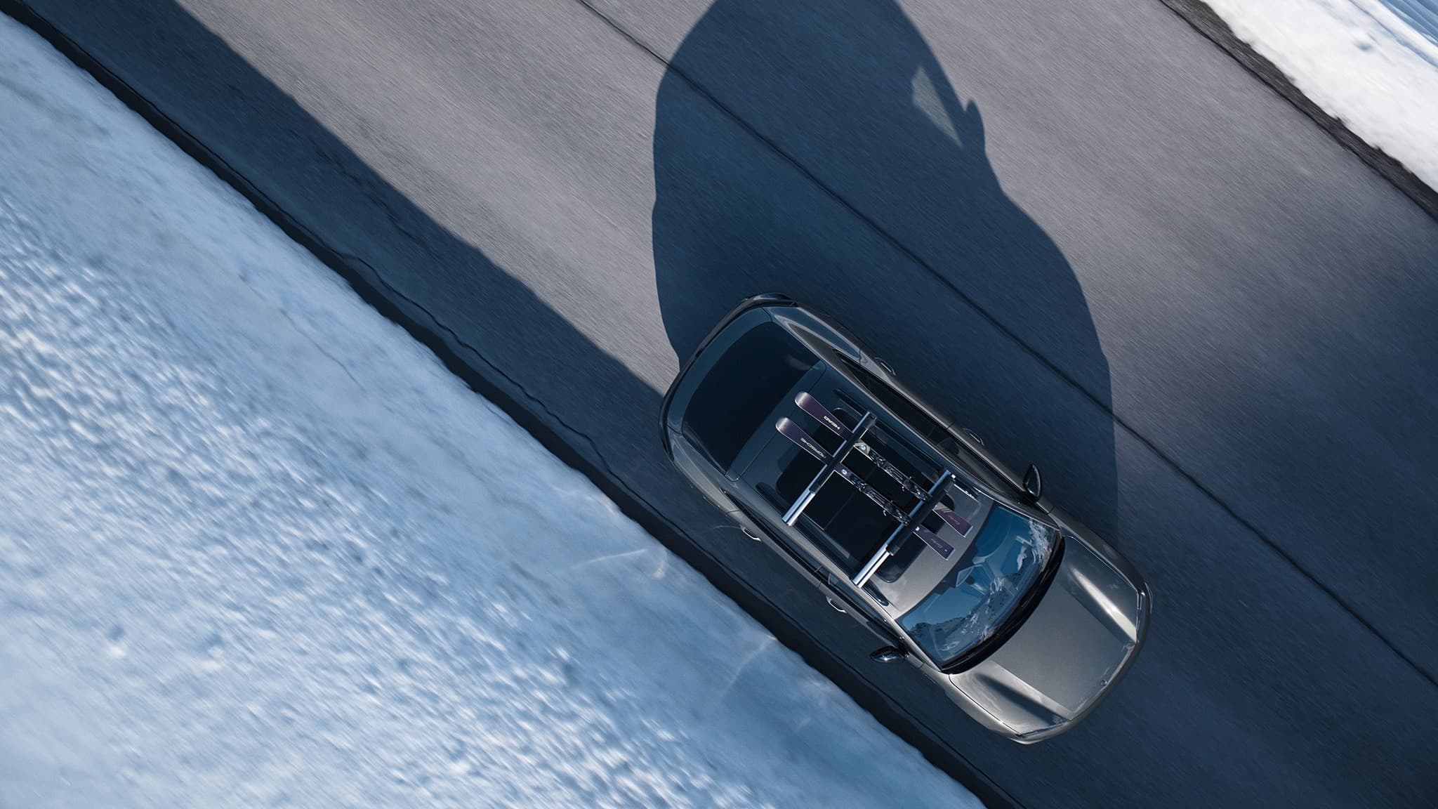Car with skis on roof driving on snowy road, aerial view, shadow cast on asphalt.