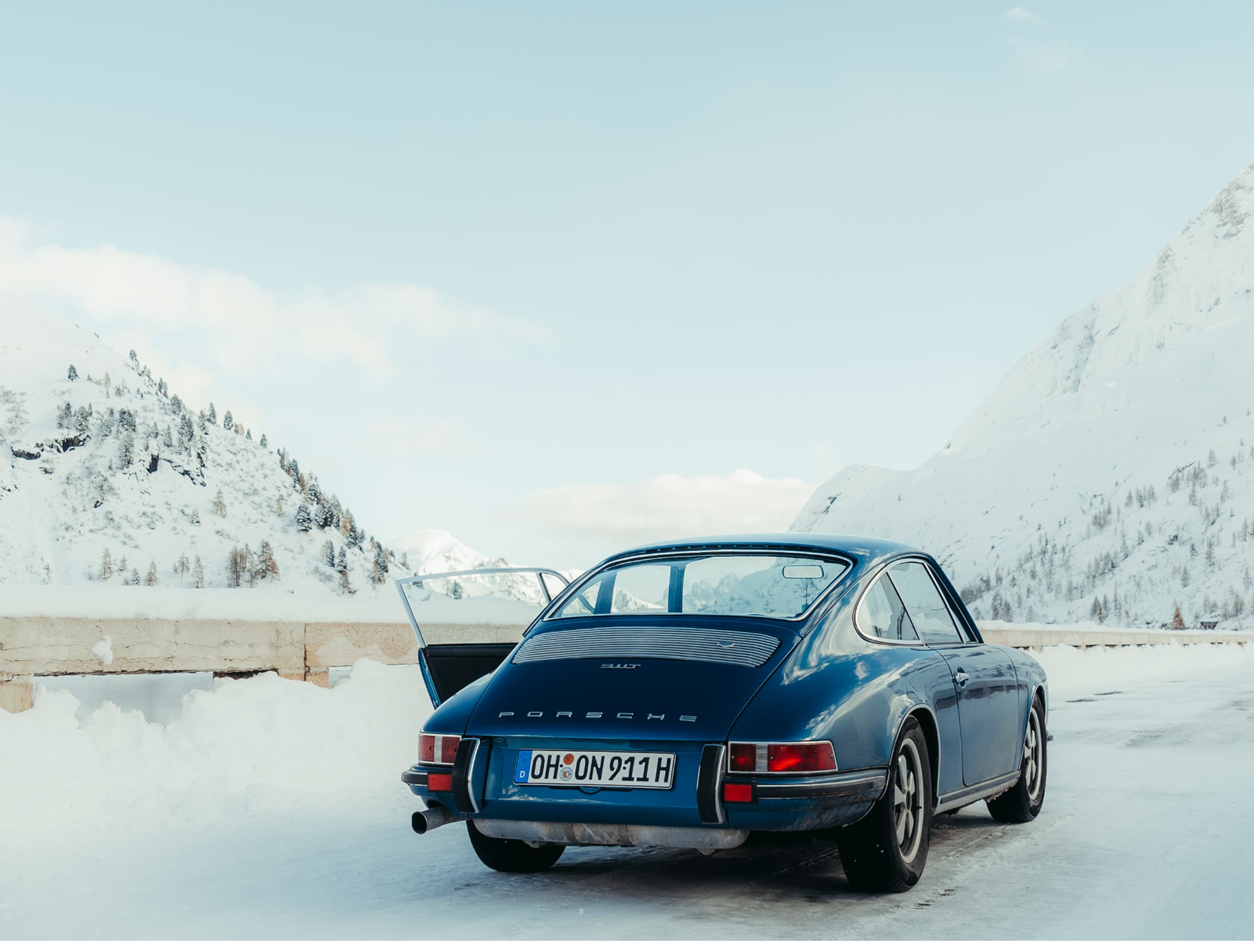 Blauer Porsche 911, geparkt auf verschneiter Bergstraße, offene Tür, klarer Himmel, schneebedeckte Gipfel im Hintergrund.