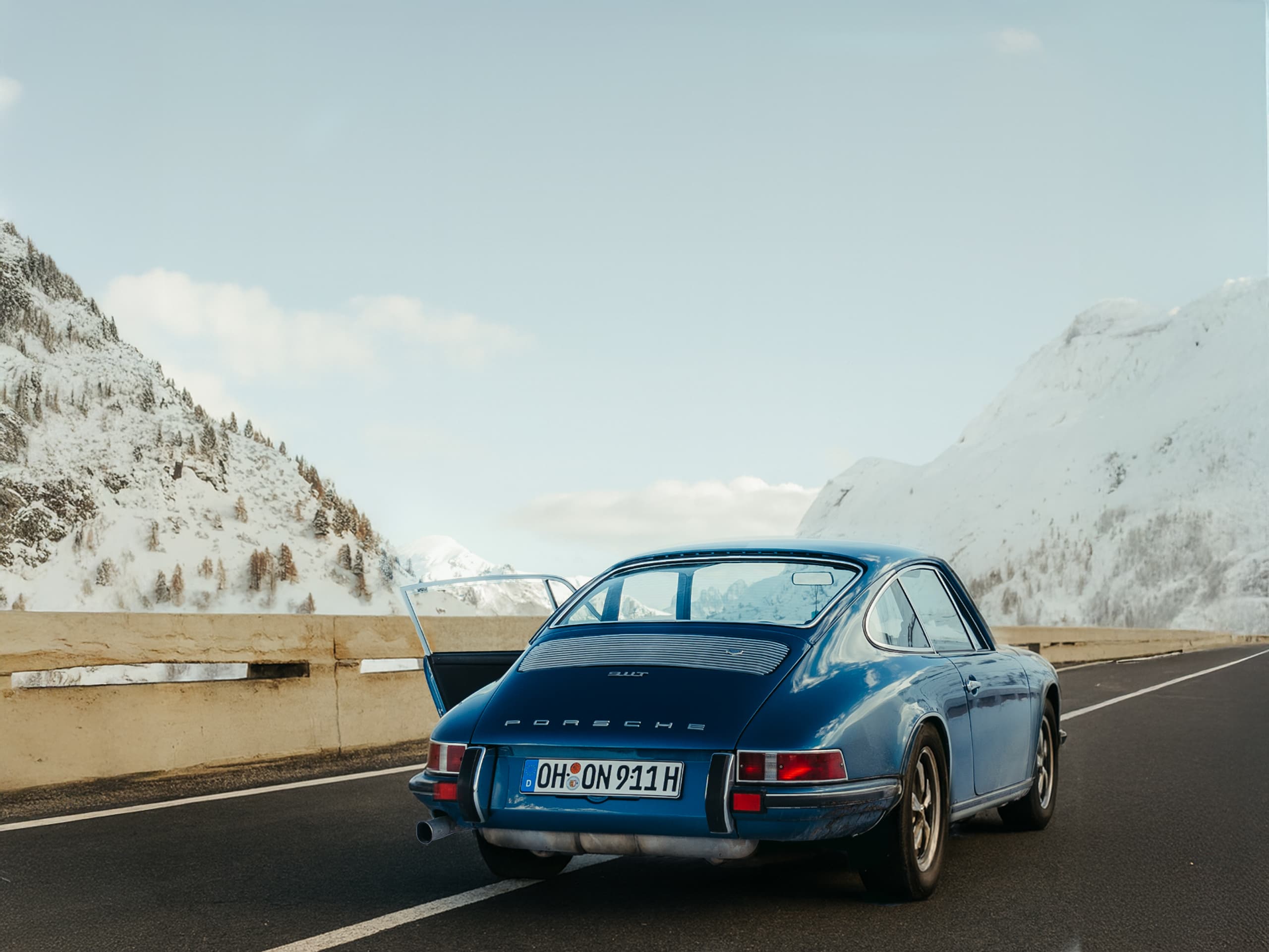 Blue Porsche 911 parked on snowy mountain road, open door, clear sky, snow-covered peaks in background.