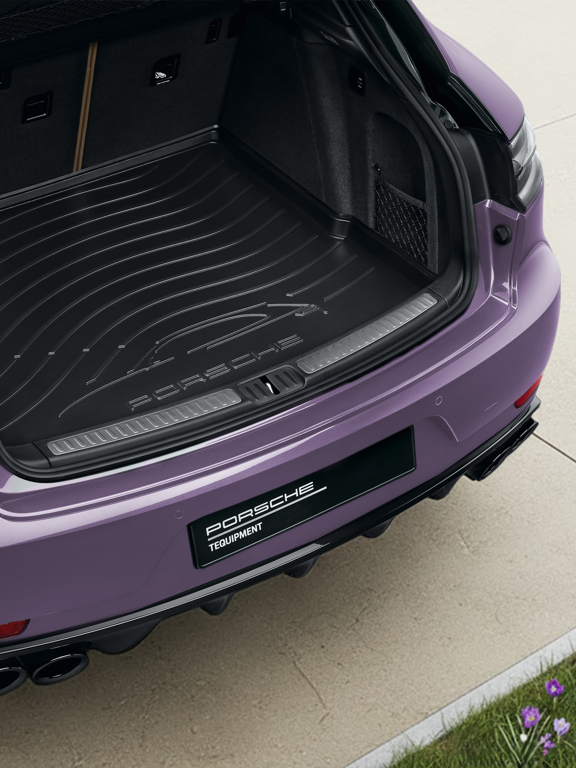 Offener Kofferraum eines violetten Porsche mit schwarzer Kofferraumauskleidung, auf einer hellen Steineinfahrt mit Gras und violetten Blumen in der Nähe.