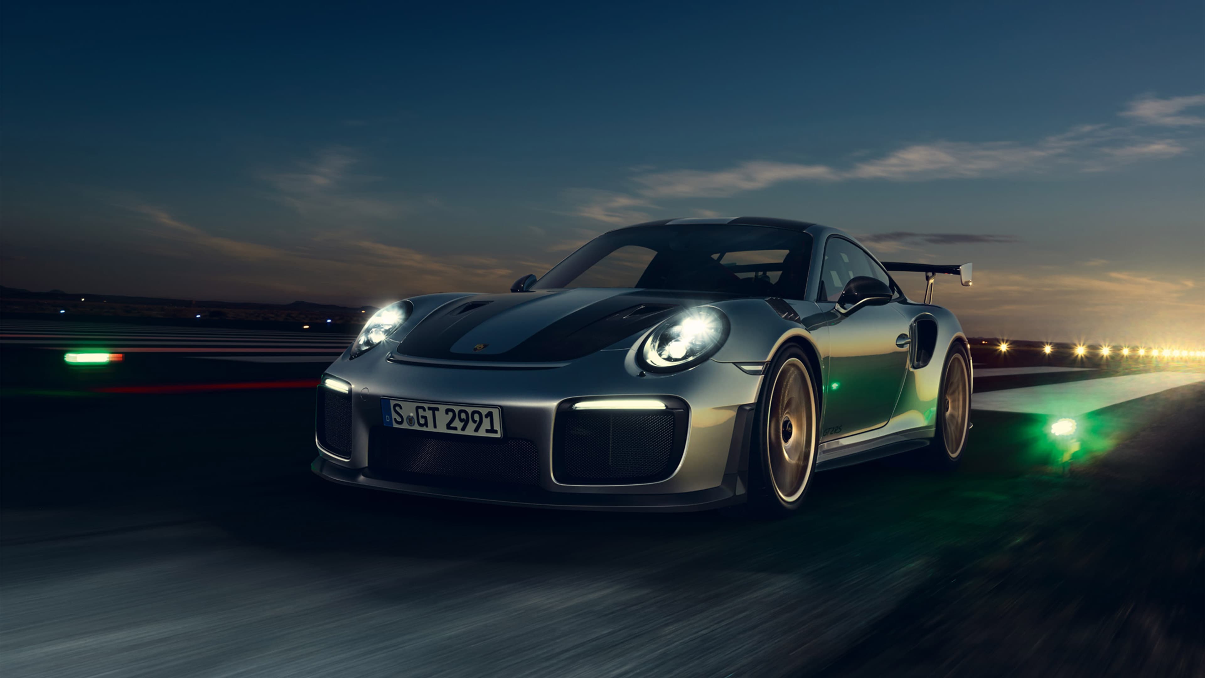 Silberner Porsche 911 GT2 RS fährt bei Dämmerung auf der Rennstrecke, Scheinwerfer an, Bewegungsunschärfe.