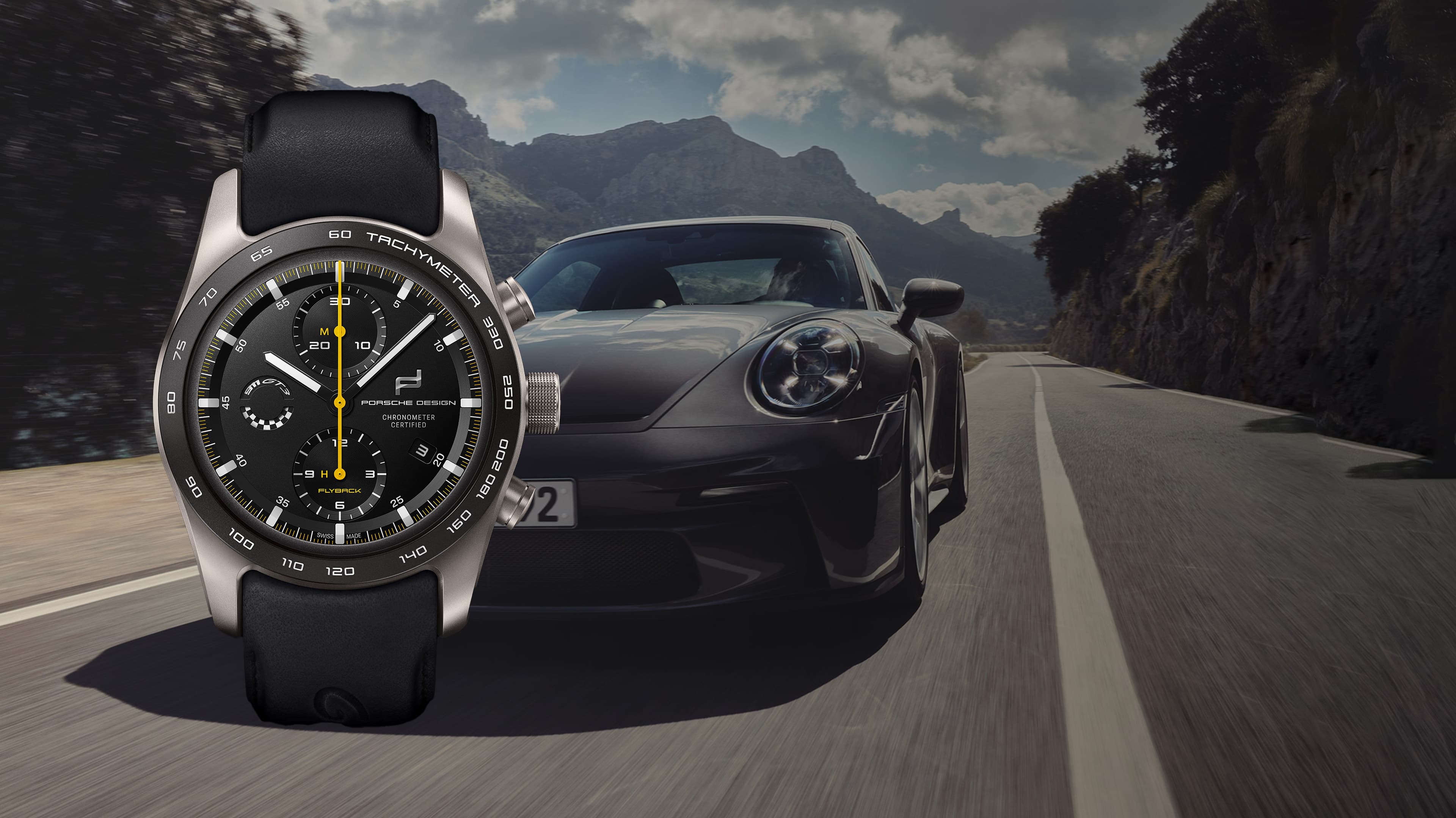 Porsche Design Chronograph mit schwarzem Armband und Tachymeter-Lünette, vor einem grauen Sportwagen auf einer Bergstraße