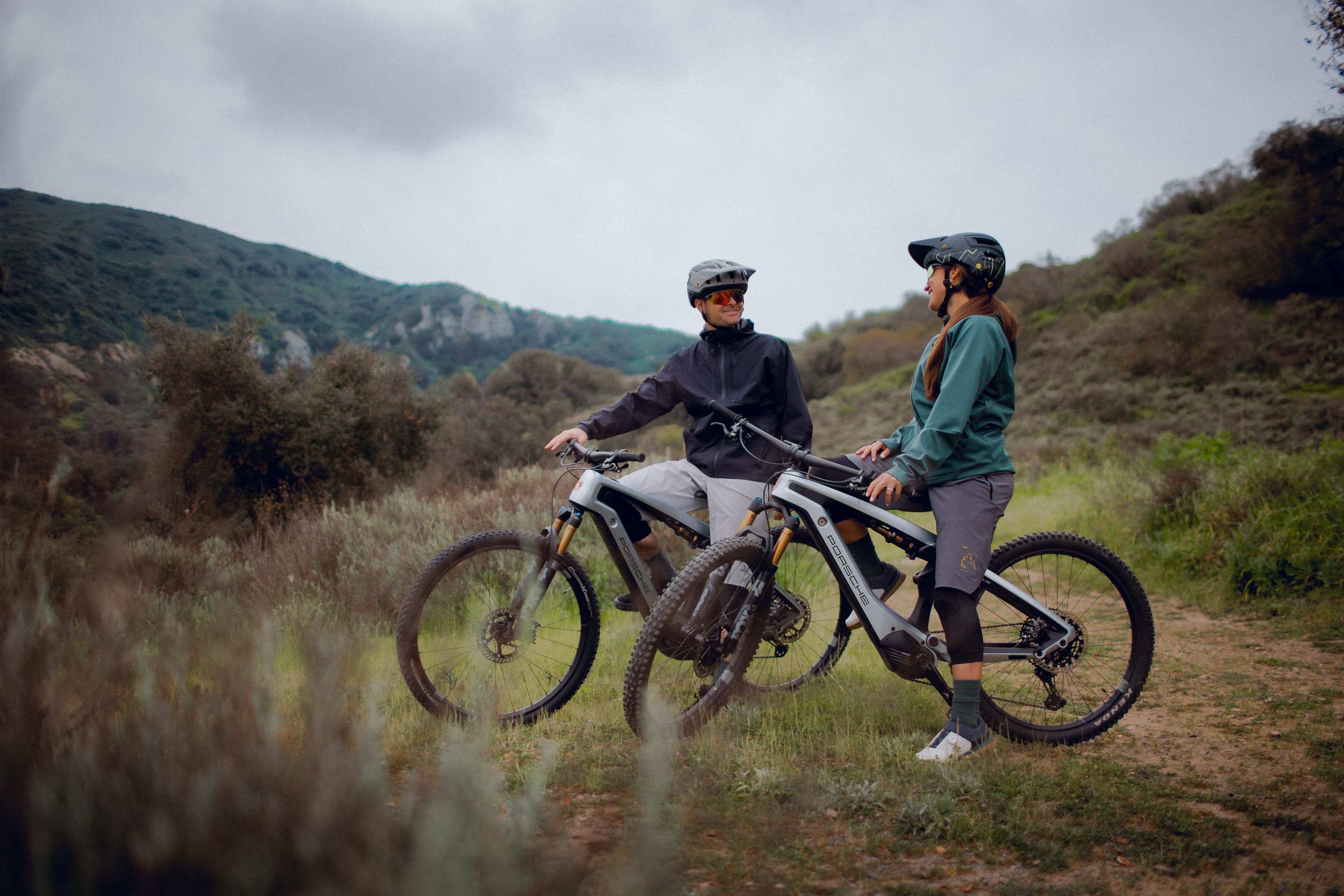 Two cyclists in helmets pause on grey e-mountain bikes on a trail, with green hills under a cloudy sky.