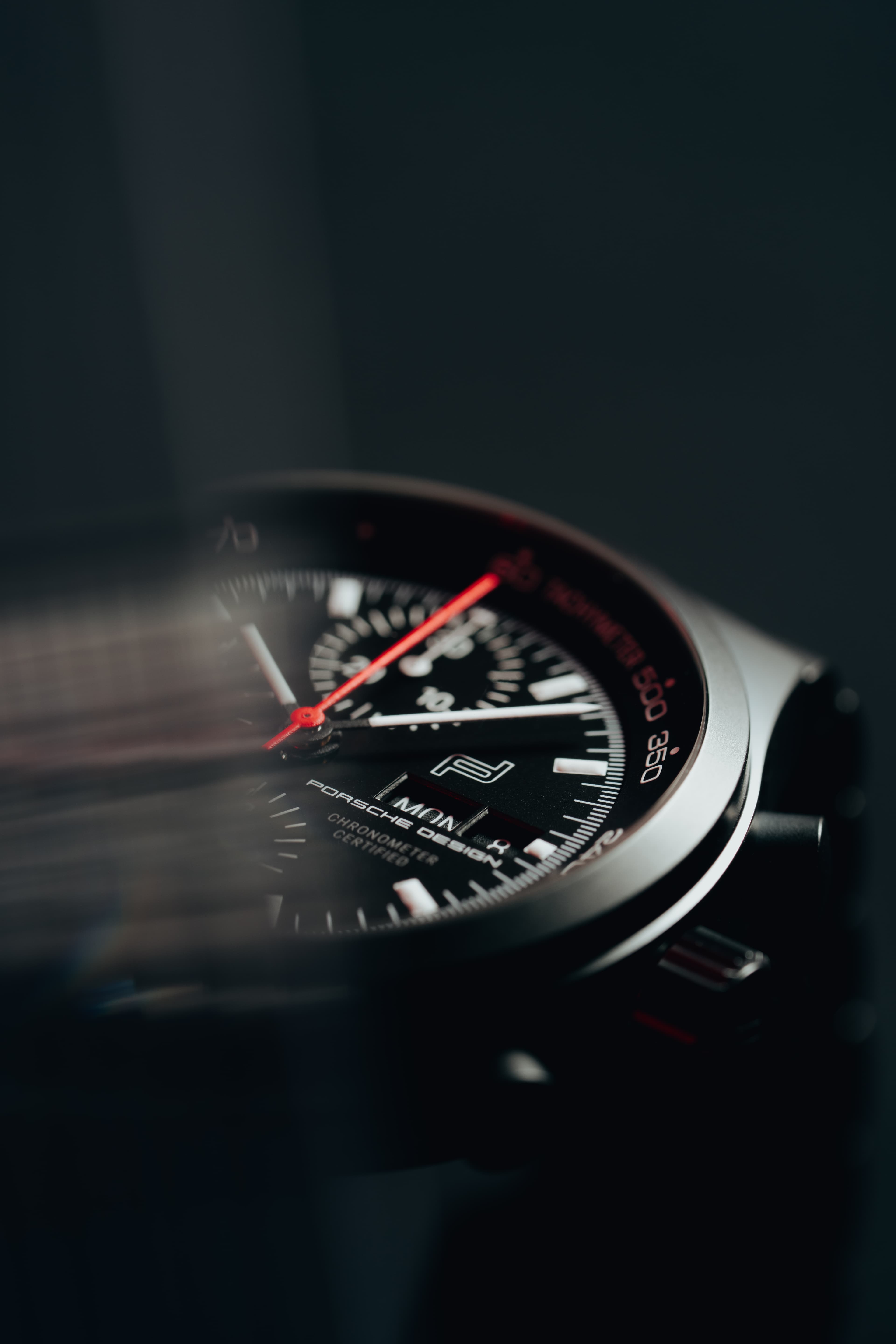 Close-up of Porsche Design chronograph watch dial with red second hand and black face in soft light.