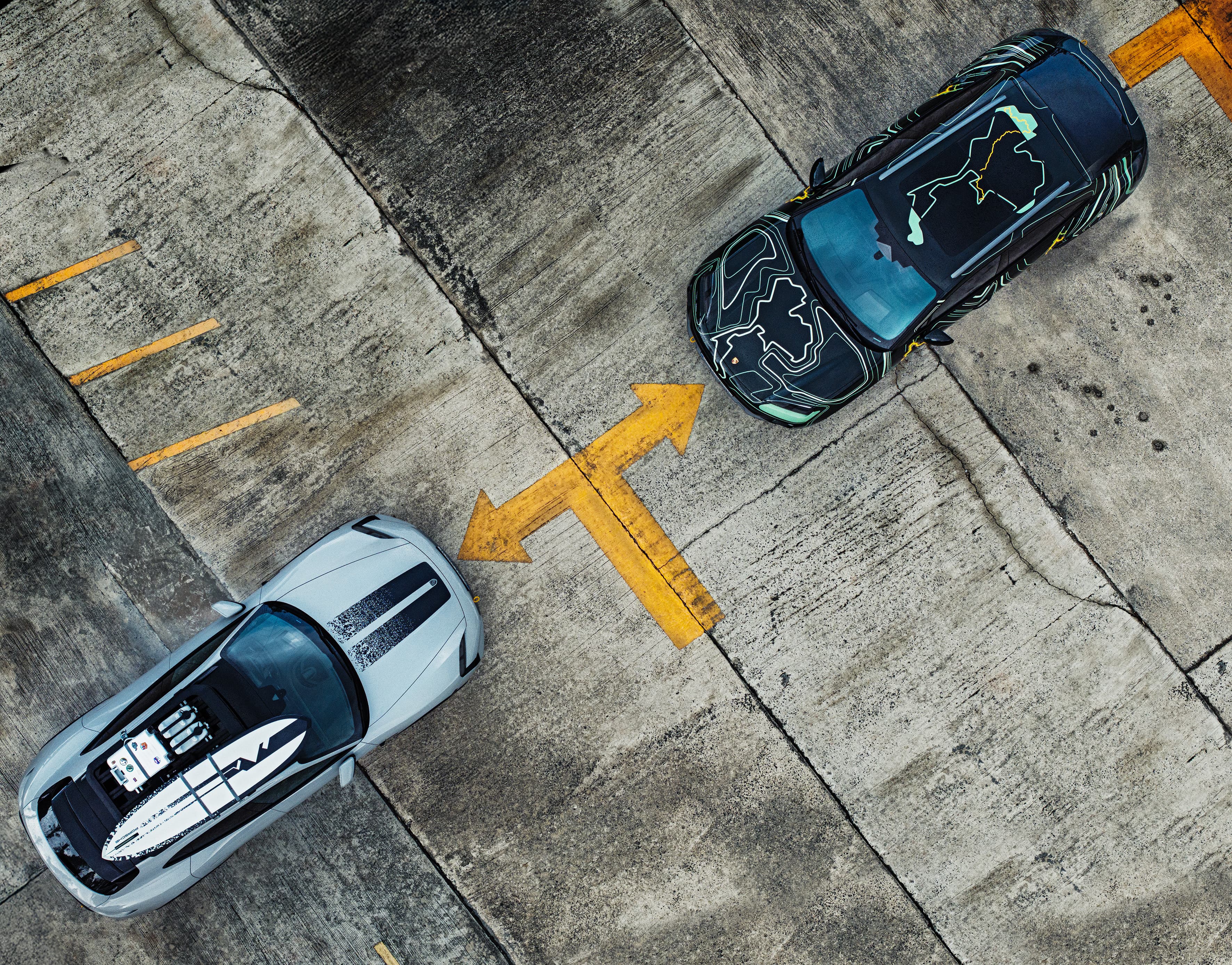 Top view: black Porsche with line camouflage wrap next to a silver Porsche on a concrete surface with a yellow arrow.