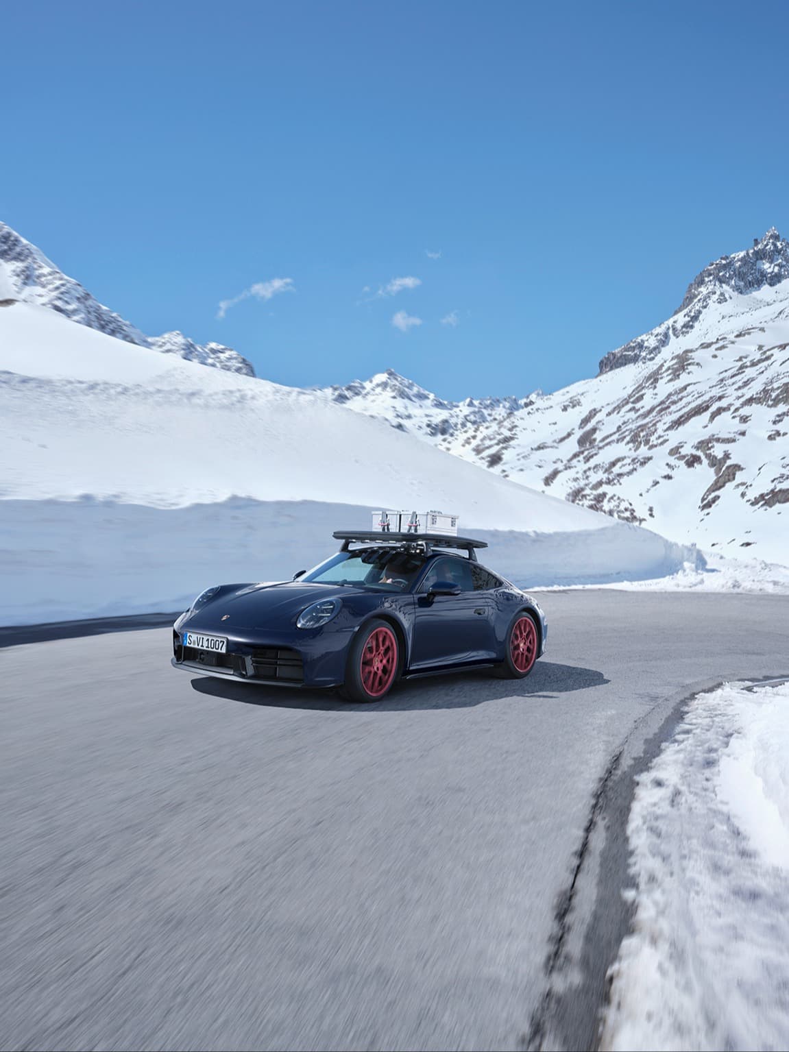 Dark blue Porsche with red wheels driving on snowy mountain road under clear blue sky. Roof rack attached.