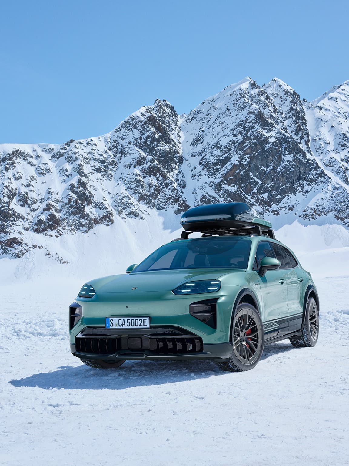 Grüner Porsche SUV mit Dachbox auf schneebedecktem Gelände, vor einer Kulisse von schneebedeckten Bergen unter klarem blauem Himmel.