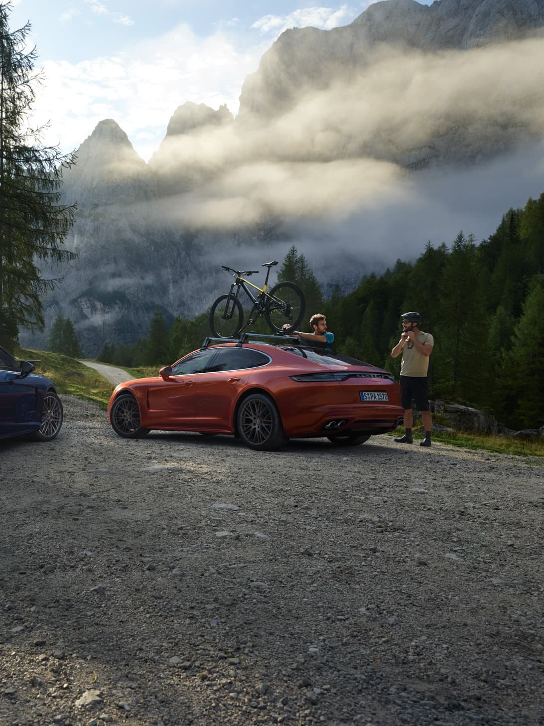 Red Porsche with a bike on the roof, parked on a mountain road, two people nearby, misty peaks in the background.