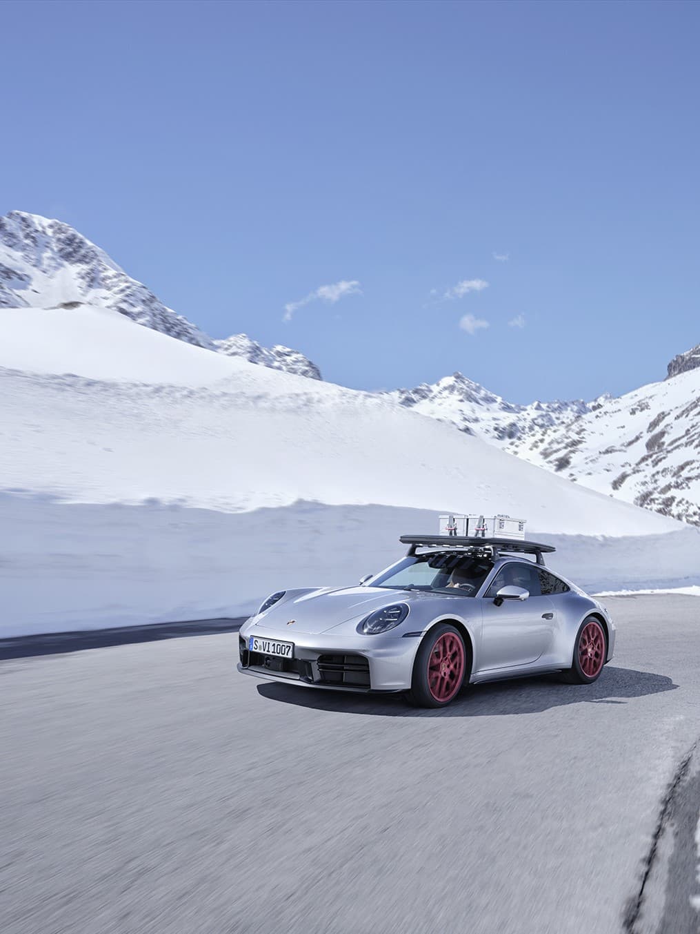 Silver Porsche with red wheels driving on snowy mountain road under clear blue sky.