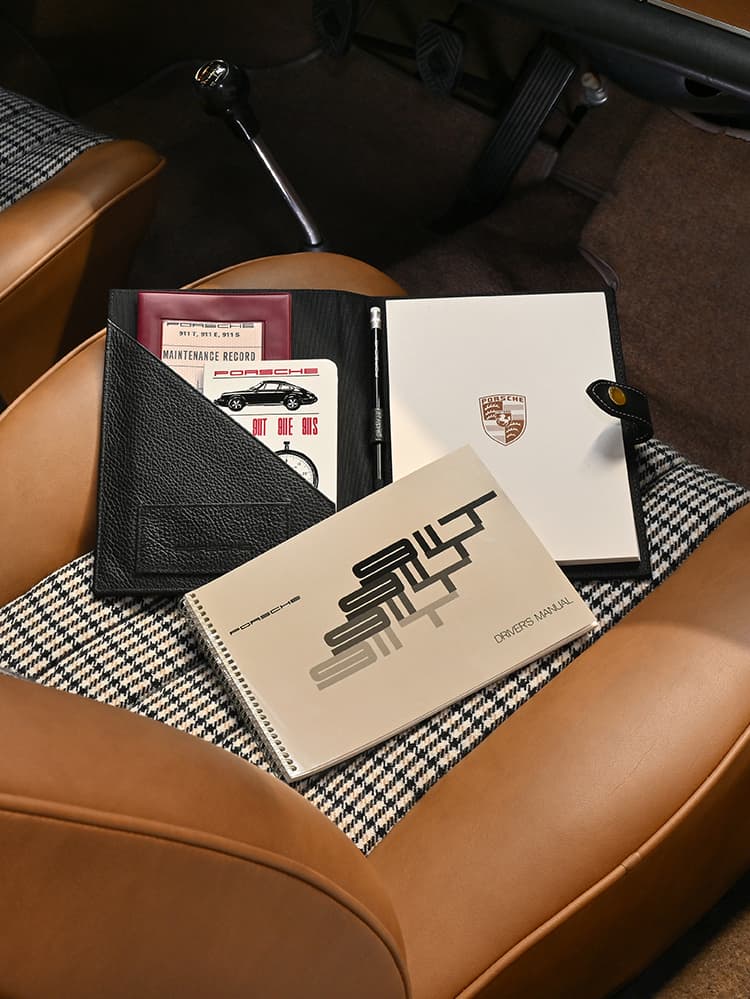 Porsche manuals and documents on houndstooth car seat, featuring black leather folder and white notebook with logo.
