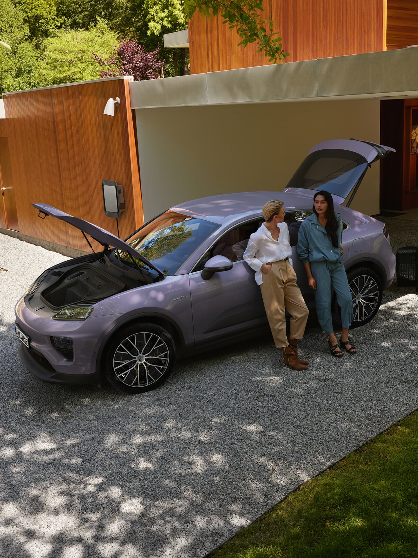 Twee vrouwen leunen tegen een paarse Porsche SUV met de motorkap vooraan en de achterklep open, voor een moderne woning.