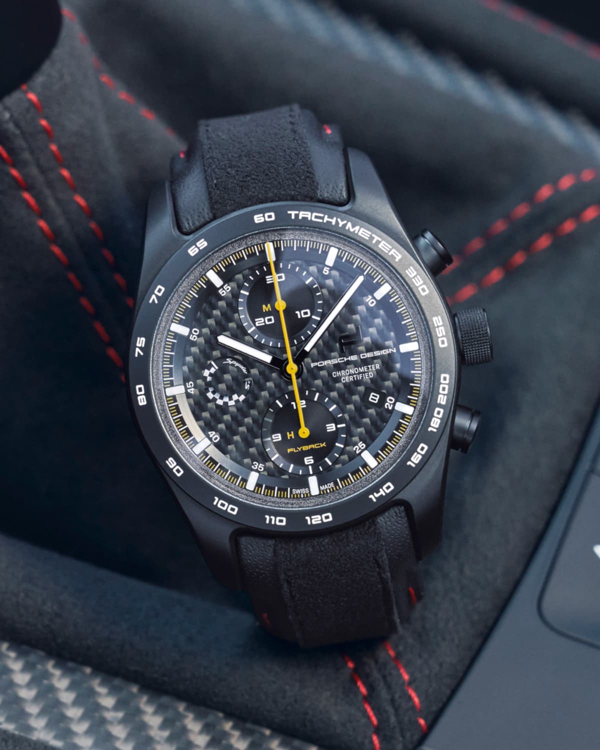 Czarny chronograf Porsche Design z tarczą z włókna węglowego i żółtą wskazówką sekundową, na tle fotela samochodowego z czerwonym przeszyciem.