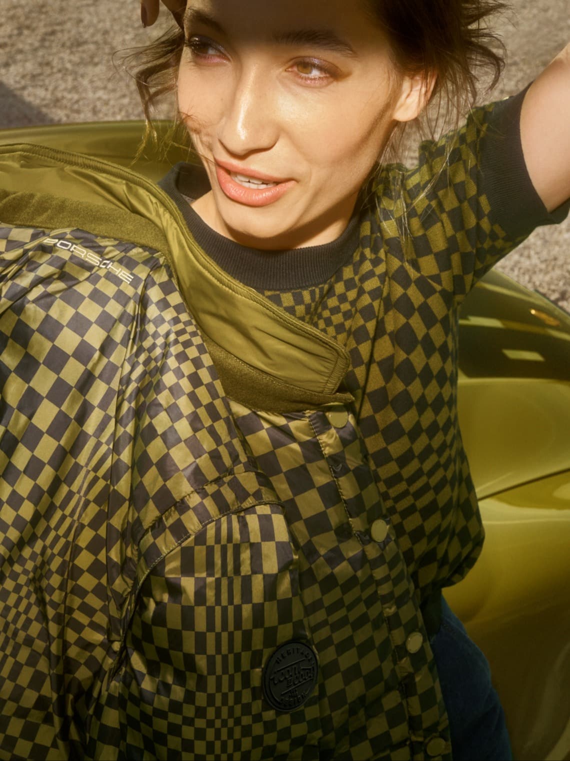 Femme portant une veste matelassée Porsche olive et noire à motif à carreaux, assise sur un siège de voiture vert, éclairée par le soleil.