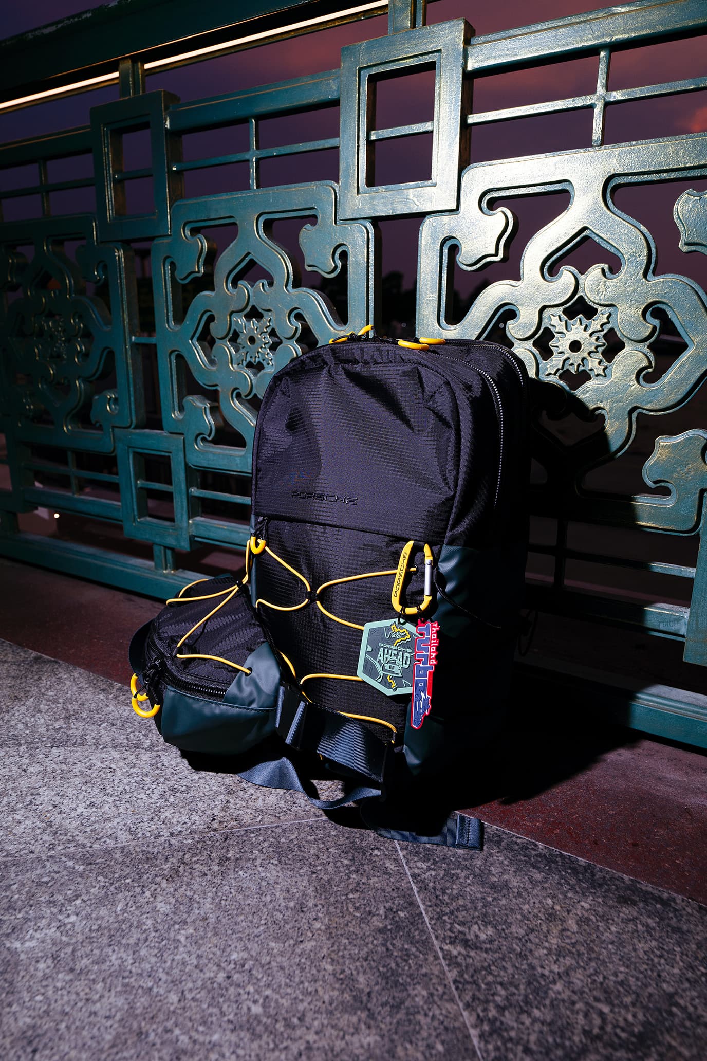Schwarzer Rucksack aus der Porsche AHEAD Collection mit gelben Kordelzügen, an ein dekoratives Metallgeländer gelehnt, im Abendlicht.