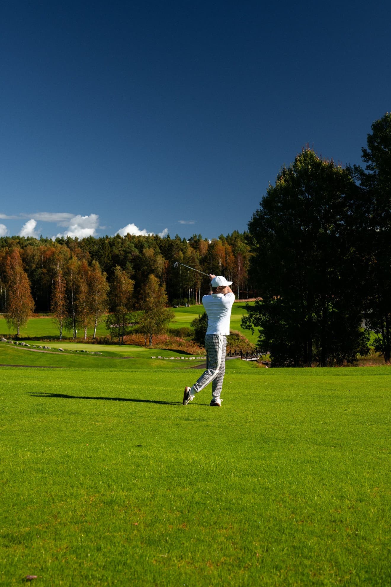 Golfer in white cap and shirt swings on green fairway, autumn trees and deep blue sky in background